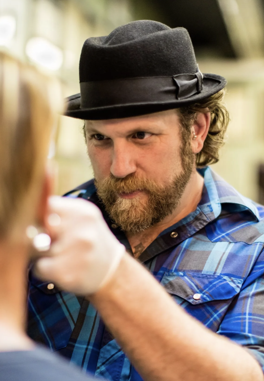 A man with a beard wearing a black fedora hat and a blue plaid shirt, looking intently at a woman elsewhere in a bar or pub setting.