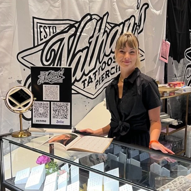 A young woman with blonde hair and a black shirt ties her waist standing behind a jewelry display counter at a tattoo shop, with a large tattoo shop sign in the background and a black sign with QR codes and payment options on the counter.