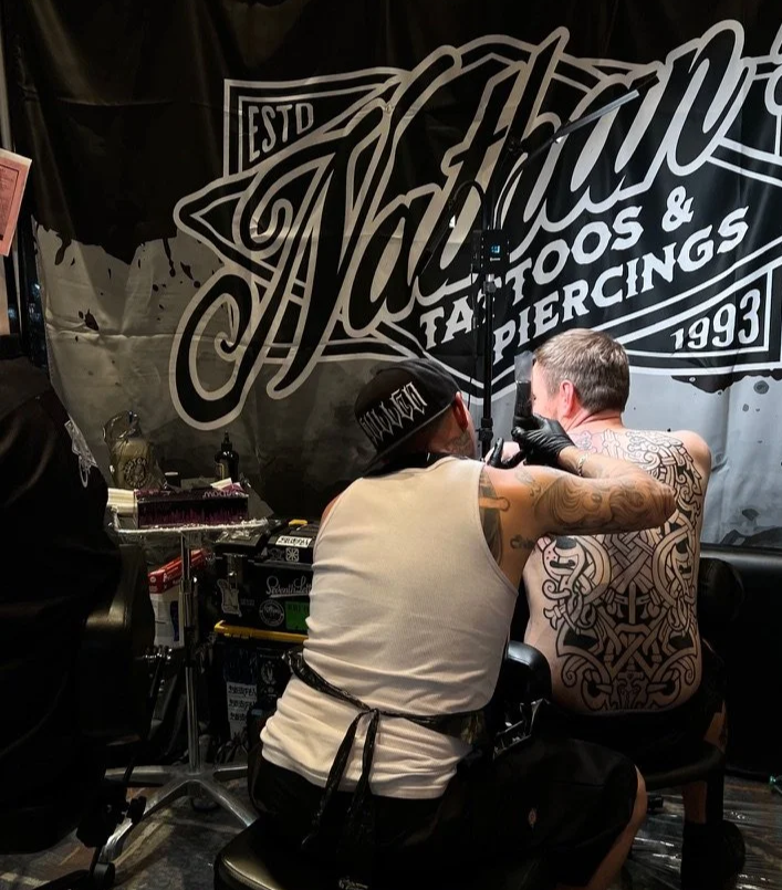 Tattoo artist working on a man's back in a tattoo shop with a banner reading 'Tattoo' and 'Tattoos & Piercings 1993' in the background.