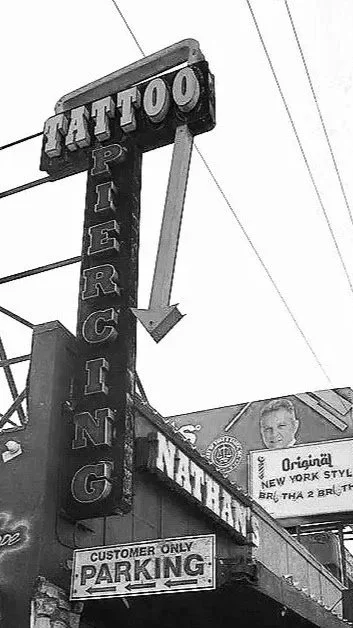 Black and white photo of a tattoo parlor sign with an arrow pointing right; signs include "Tattoo" at the top, "Piercing" vertically, and a parking sign below.