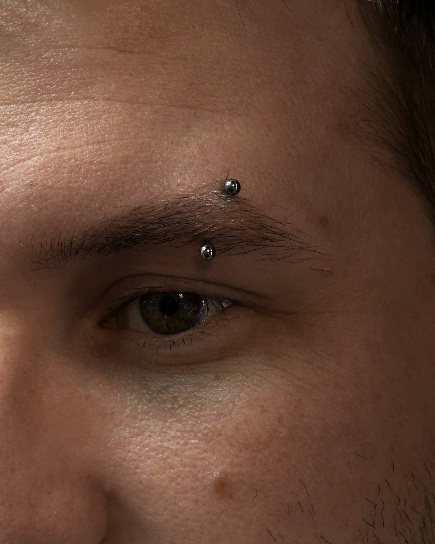 Close-up image of a person's forehead with two silver studs pierced through the skin above the eyebrow.