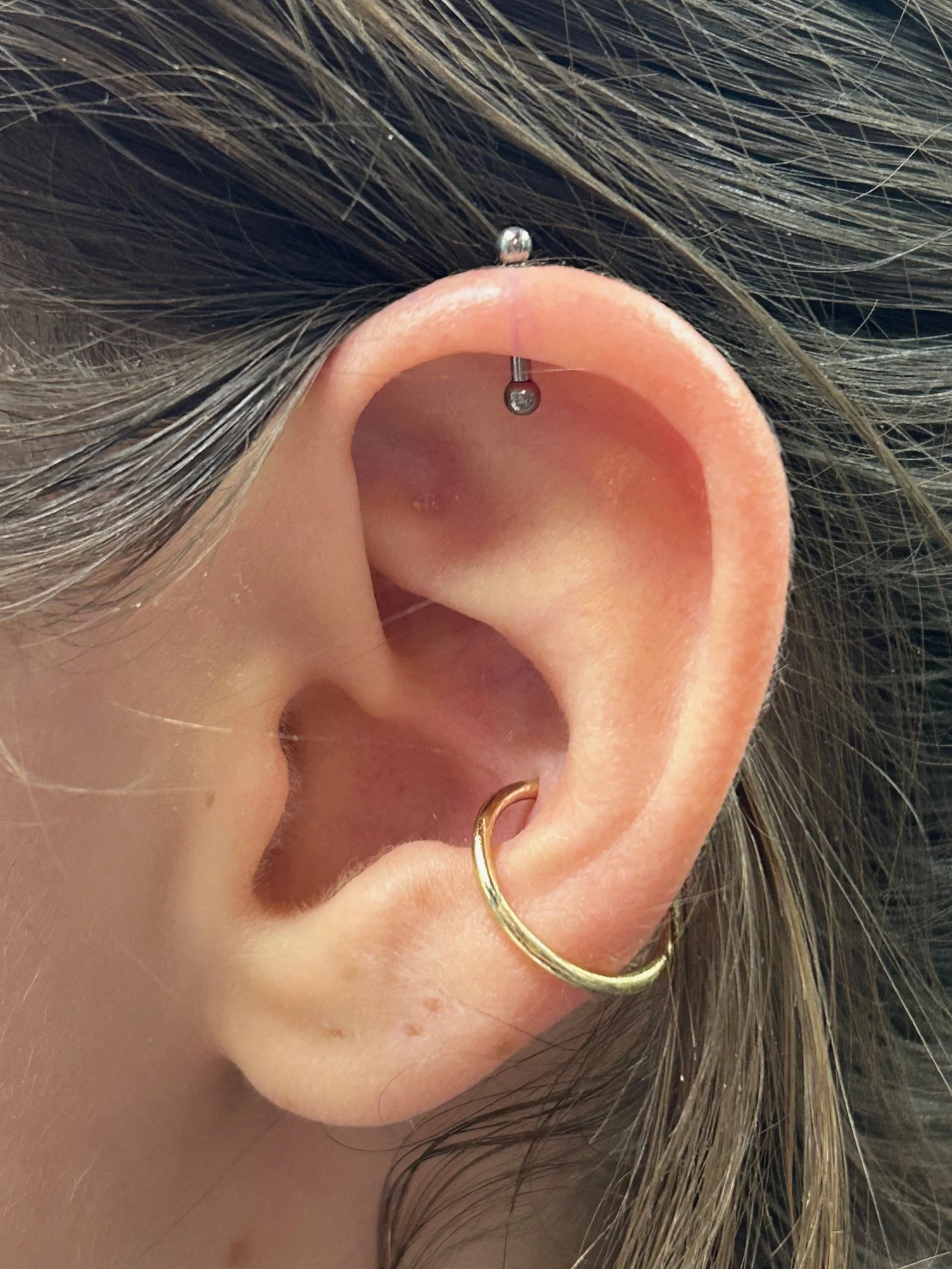 Close-up of a person's ear with a small black stud earring on the top part of the ear and a gold hoop earring on the earlobe, with brown and blonde hair surrounding the ear.