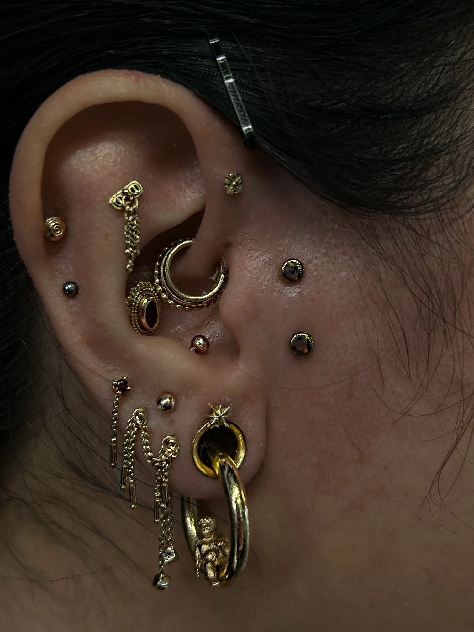 Close-up of a person's ear with several gold and black jewelry pieces, including earrings, ear cuffs, and chains, with hair partially covering the ear.