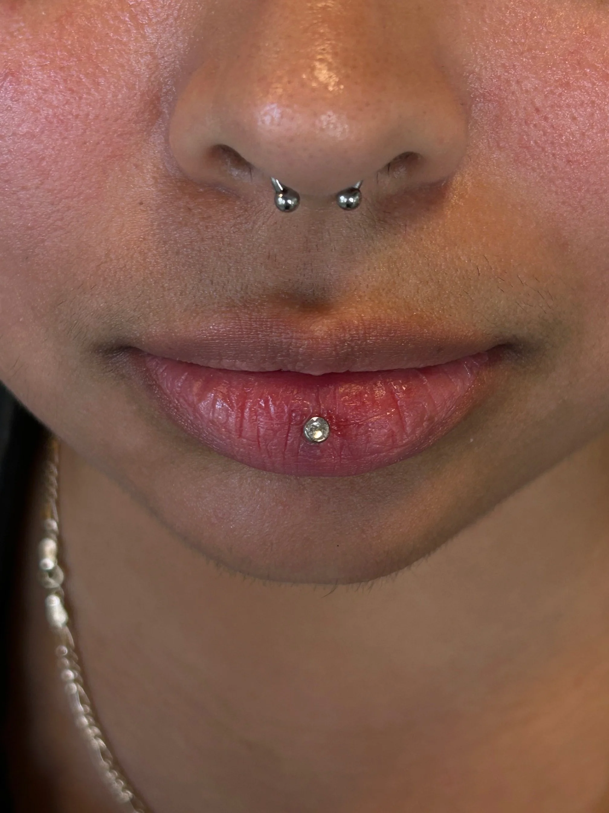 Close-up of a person's nose and lips, featuring nose piercings with metal studs and a bottom lip piercing with a metallic gem.
