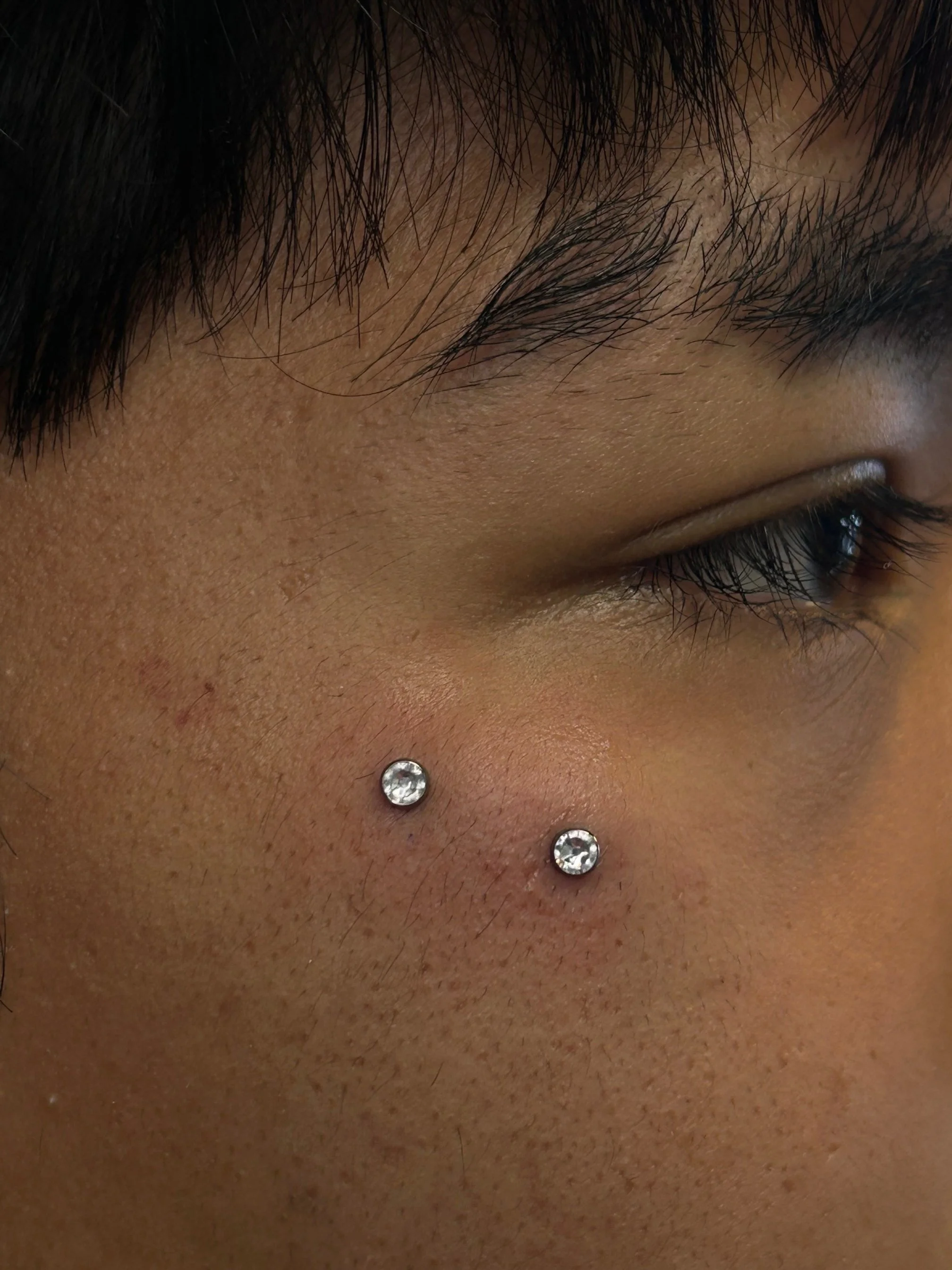 Close-up of a person's face, showing part of their eyebrow, closed eye, and two rhinestones on the lower cheek.