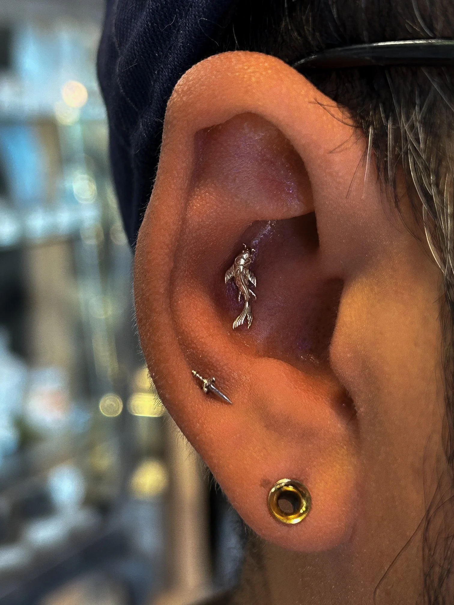 Close-up of a person's ear with multiple piercings, including a helix piercing with a silver hoop, a tragus piercing with a gold ring, and a lobe piercing with a small gold jewelry piece. The background is blurred.