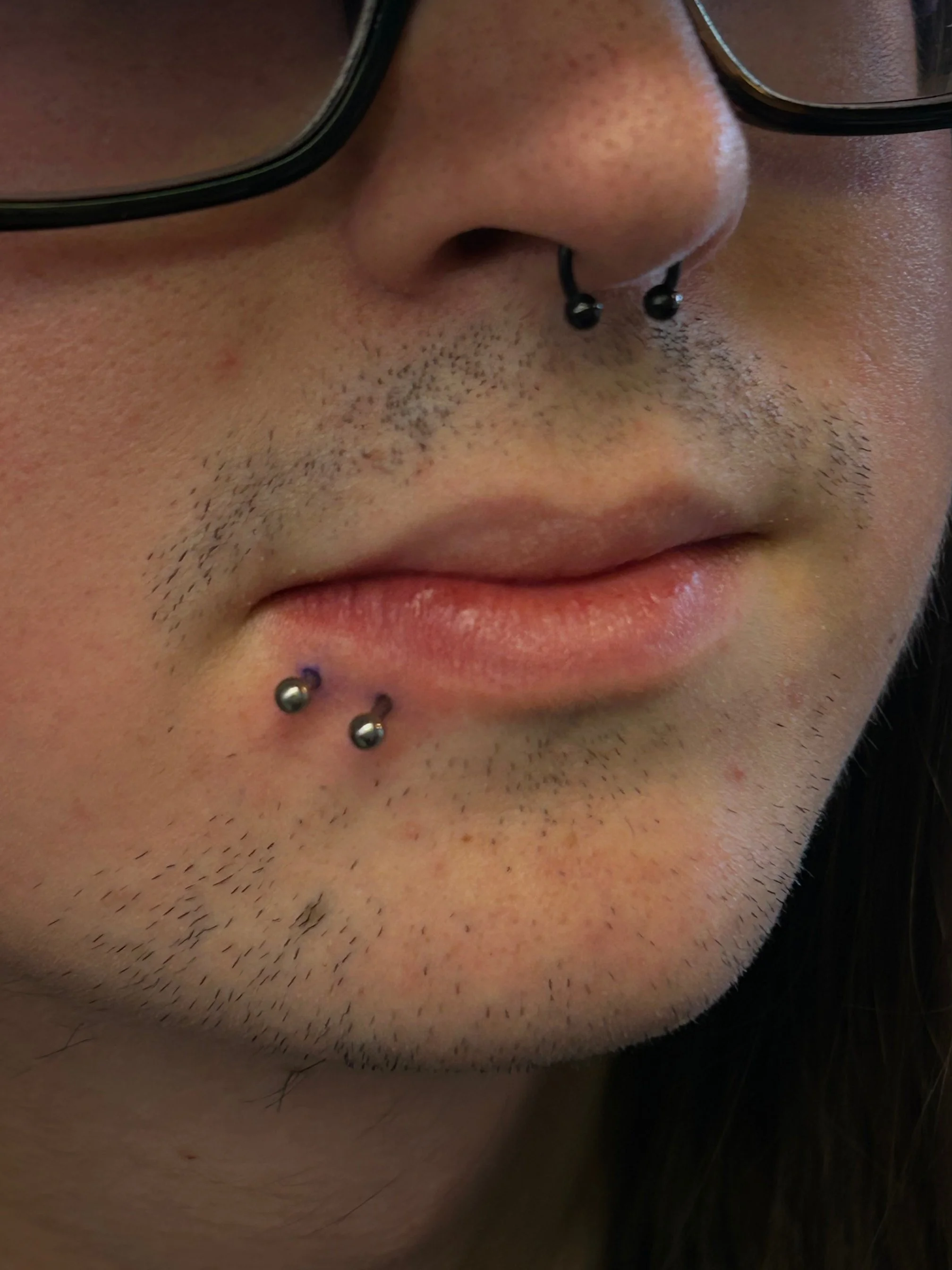Close-up of a person's face with facial hair, wearing black-rimmed glasses, and multiple facial piercings, including a septum and two piercings below the lower lip.