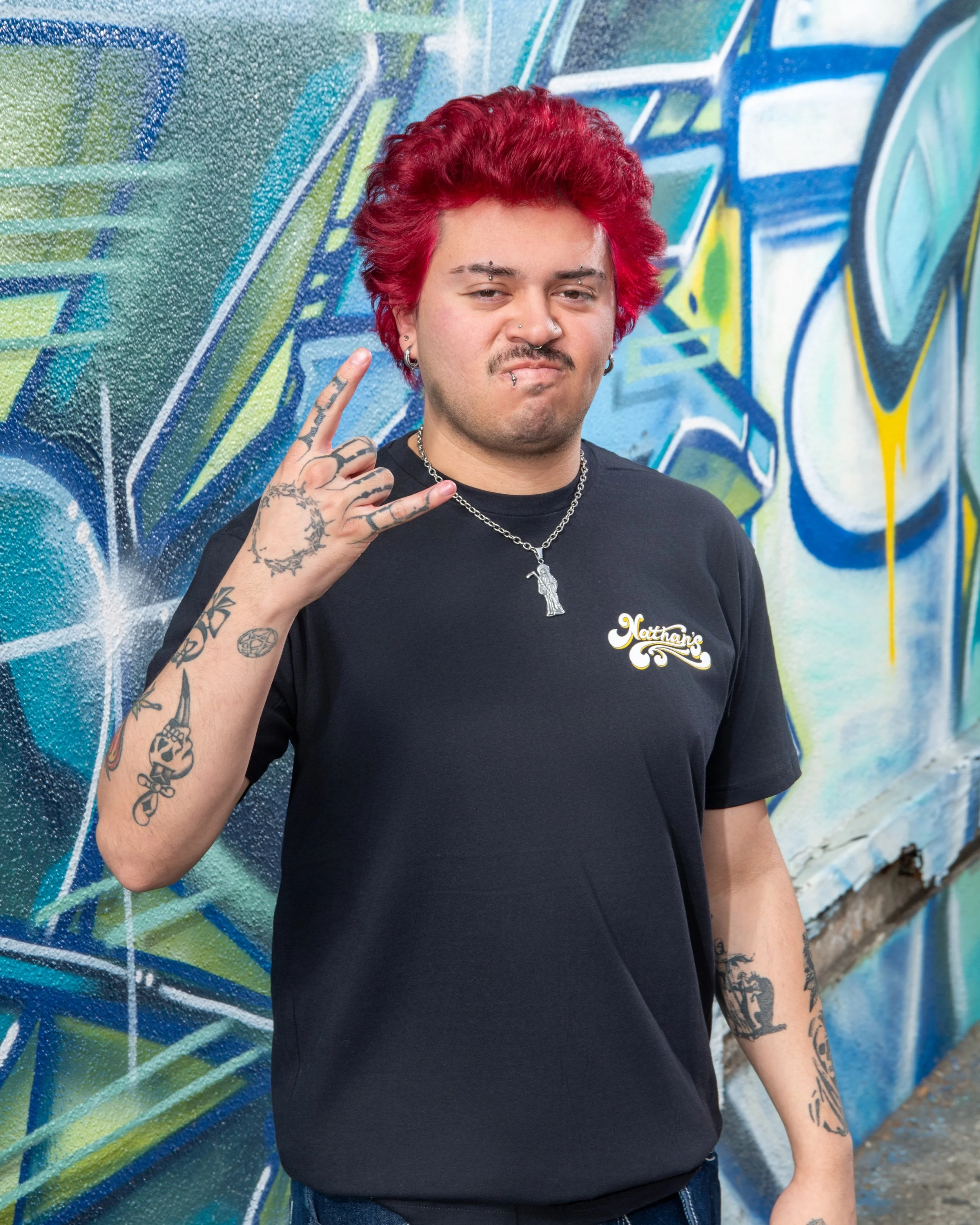 A person with red hair and multiple facial and ear piercings, tattoos on their arm and hand, wearing a black t-shirt with a logo, standing in front of a colorful graffiti wall, making a hand gesture with their ring finger and pinky extended.