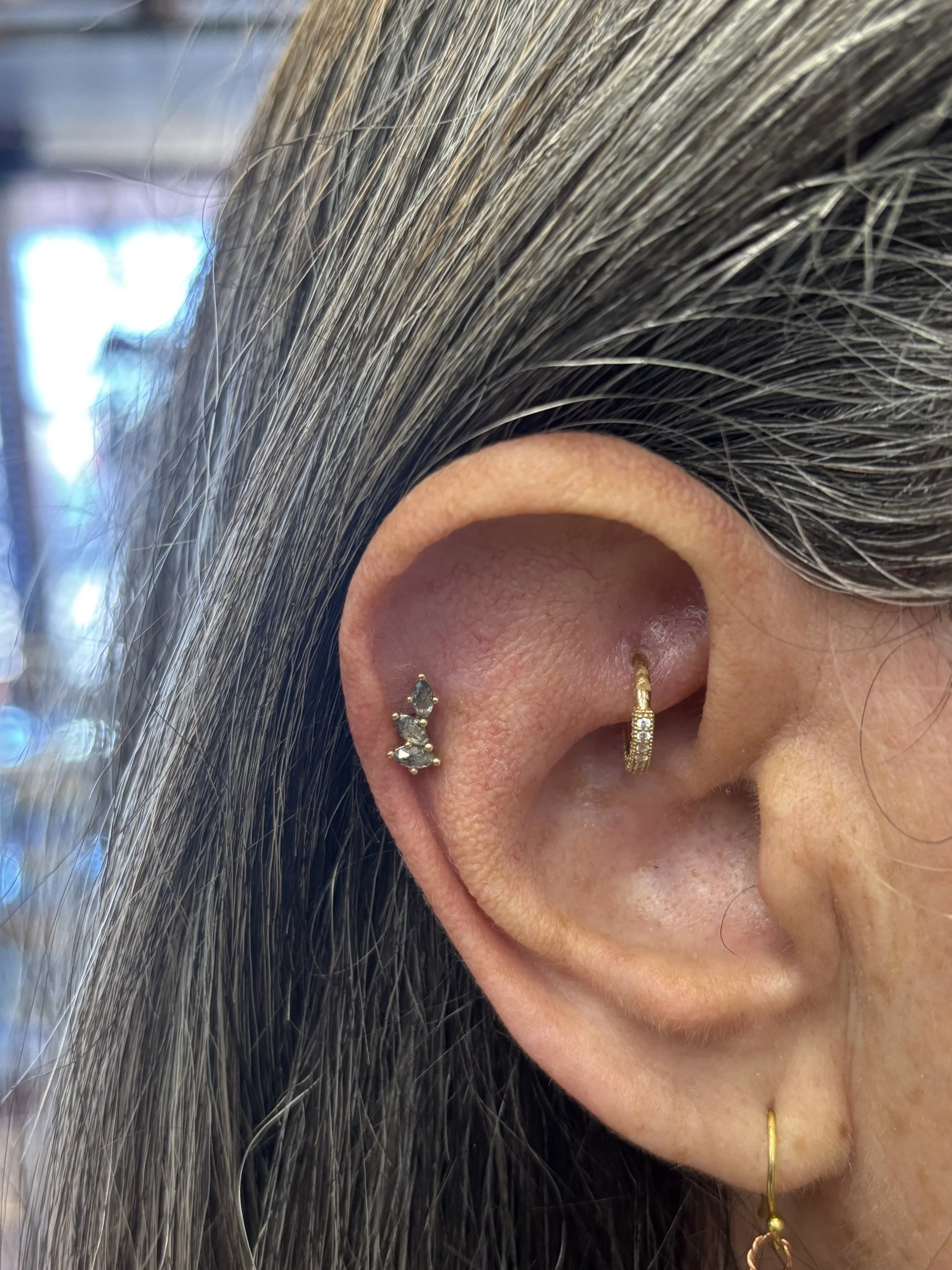 Close-up of a person's ear with multiple earrings, including a small hoop with rhinestones, a small gold hoop, and a gold dangling earring with white stones. The person has long, dark hair with gray strands.