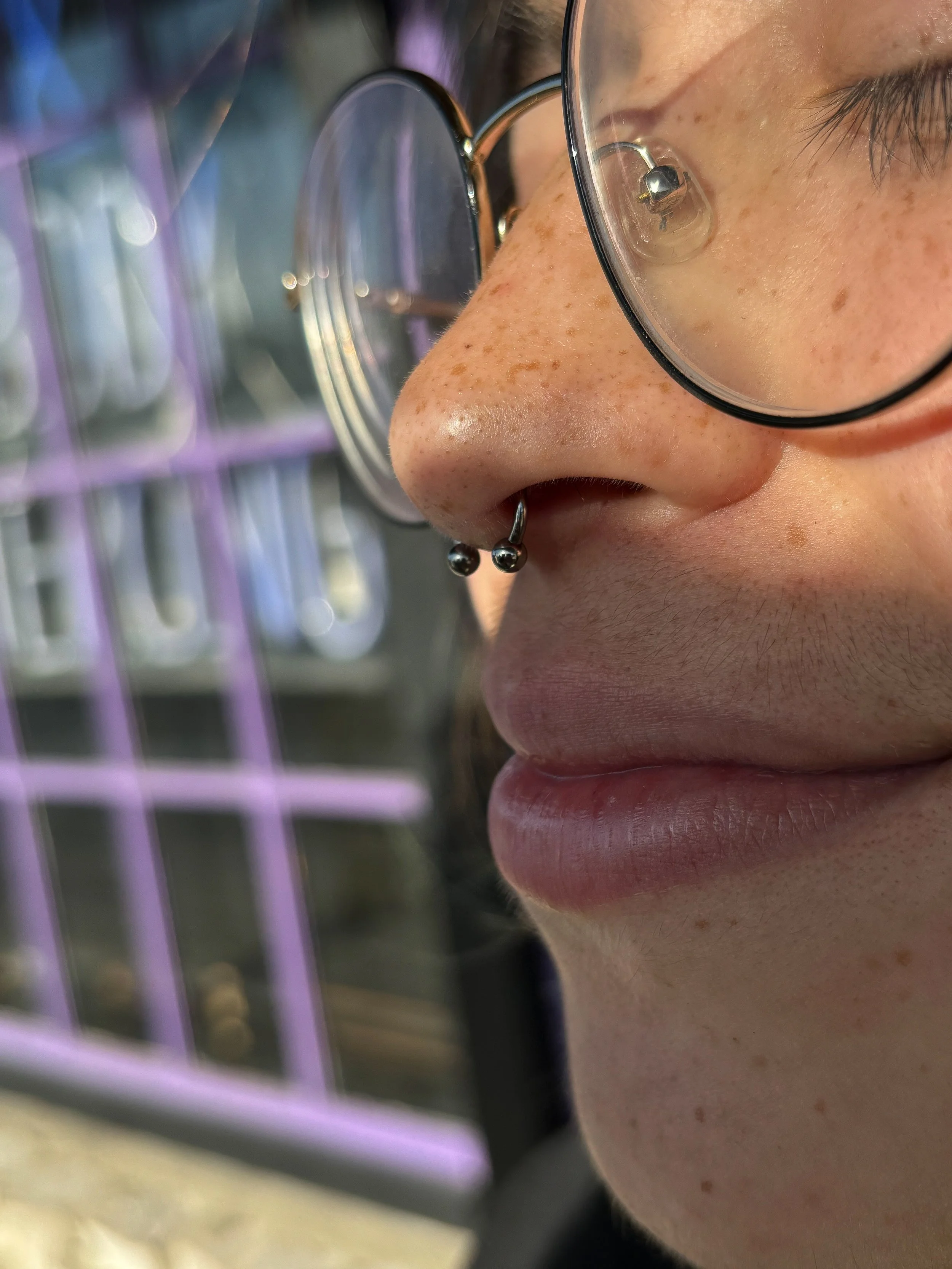 Close-up of a person's face with glasses and piercing, with a purple clock in the background.