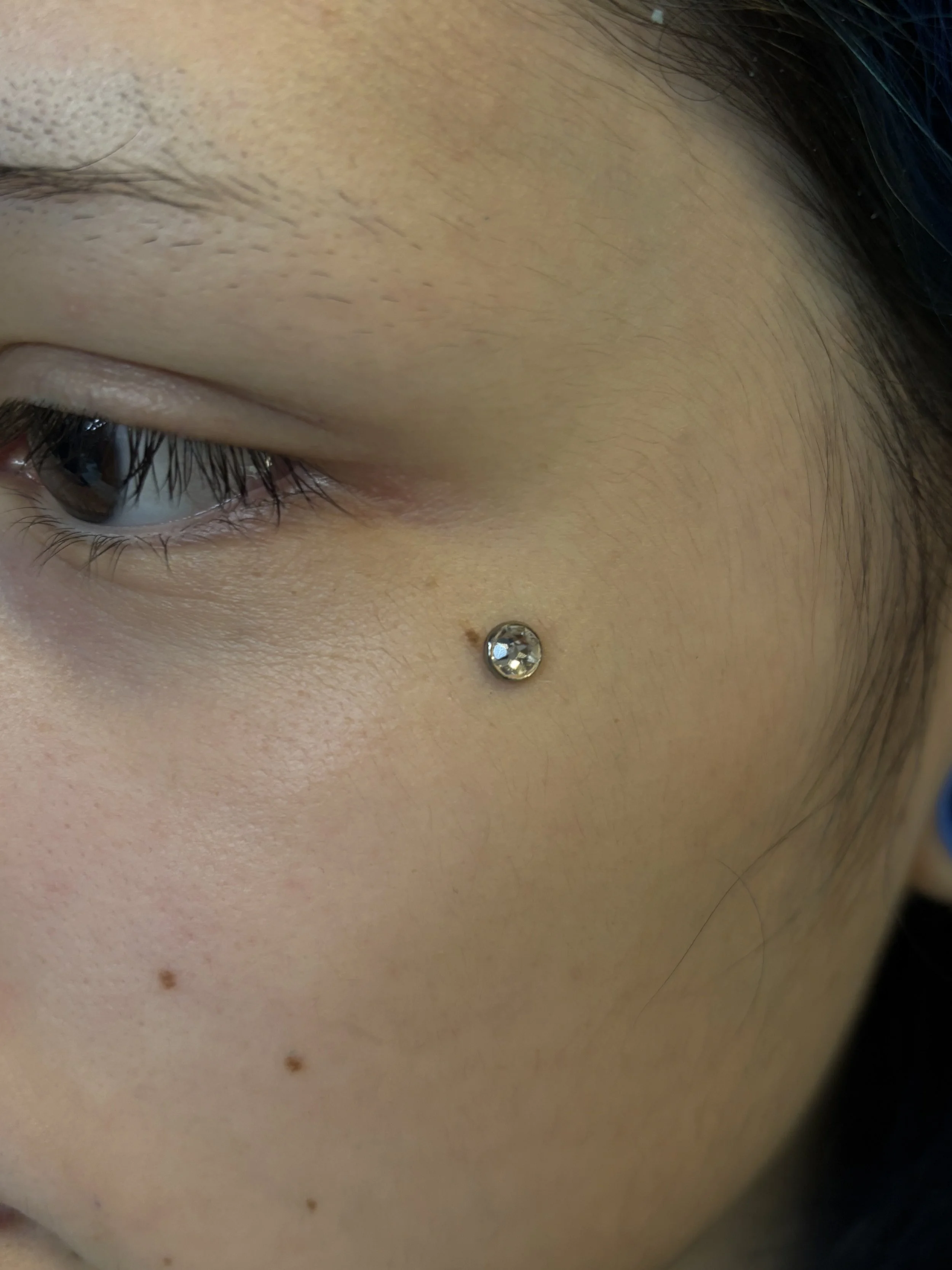 Close-up of a person's eye, showing eyelashes, skin texture, and a small metallic stud piercing below the eye.