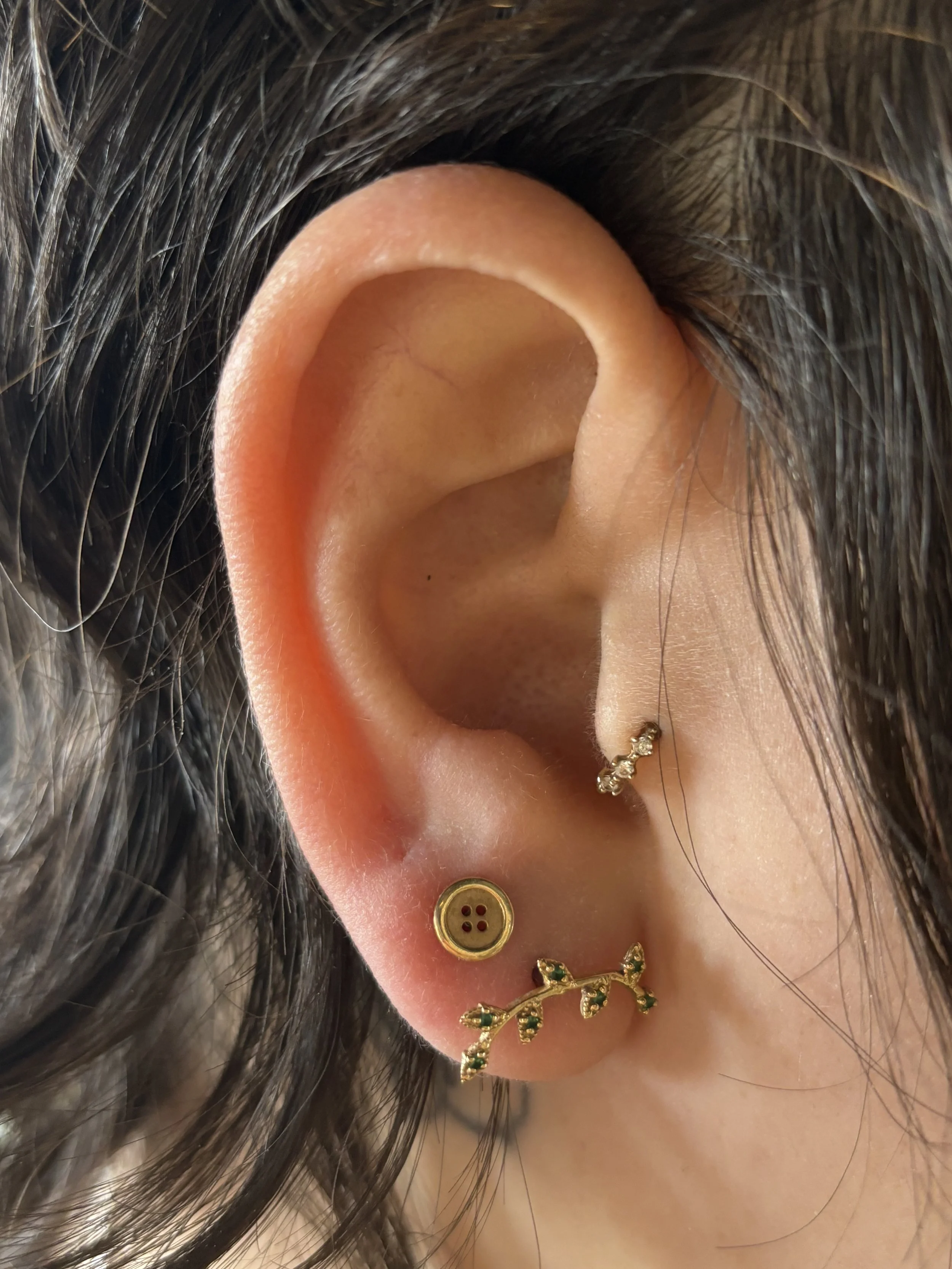 Close-up of a person's ear with three earrings: a gold button ear cuff, a gold and green leaf-shaped earring, and a small gold hoop with a dangling charm.