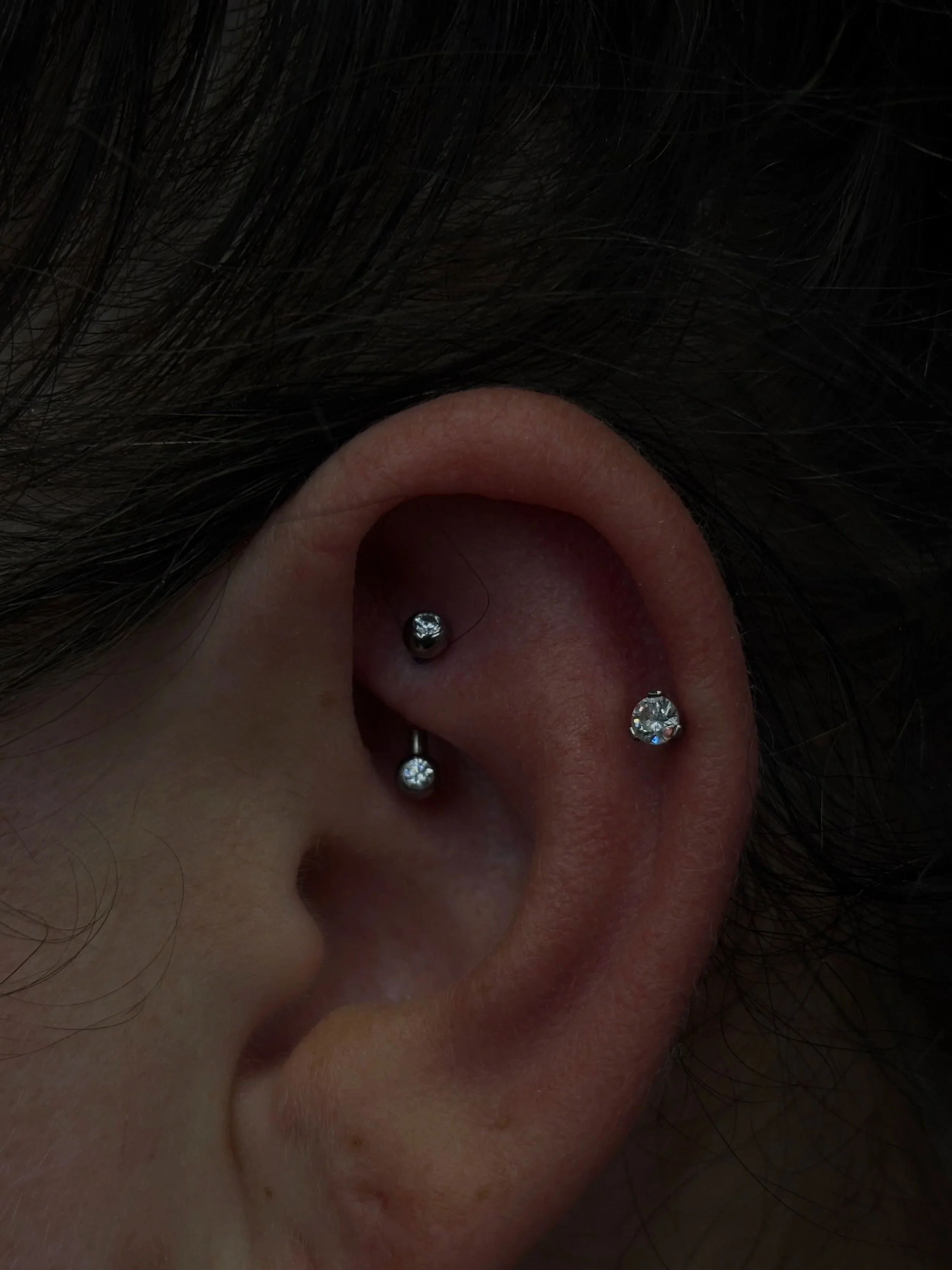 Close-up of a person's ear with multiple piercings and a nose screw, surrounded by dark hair.