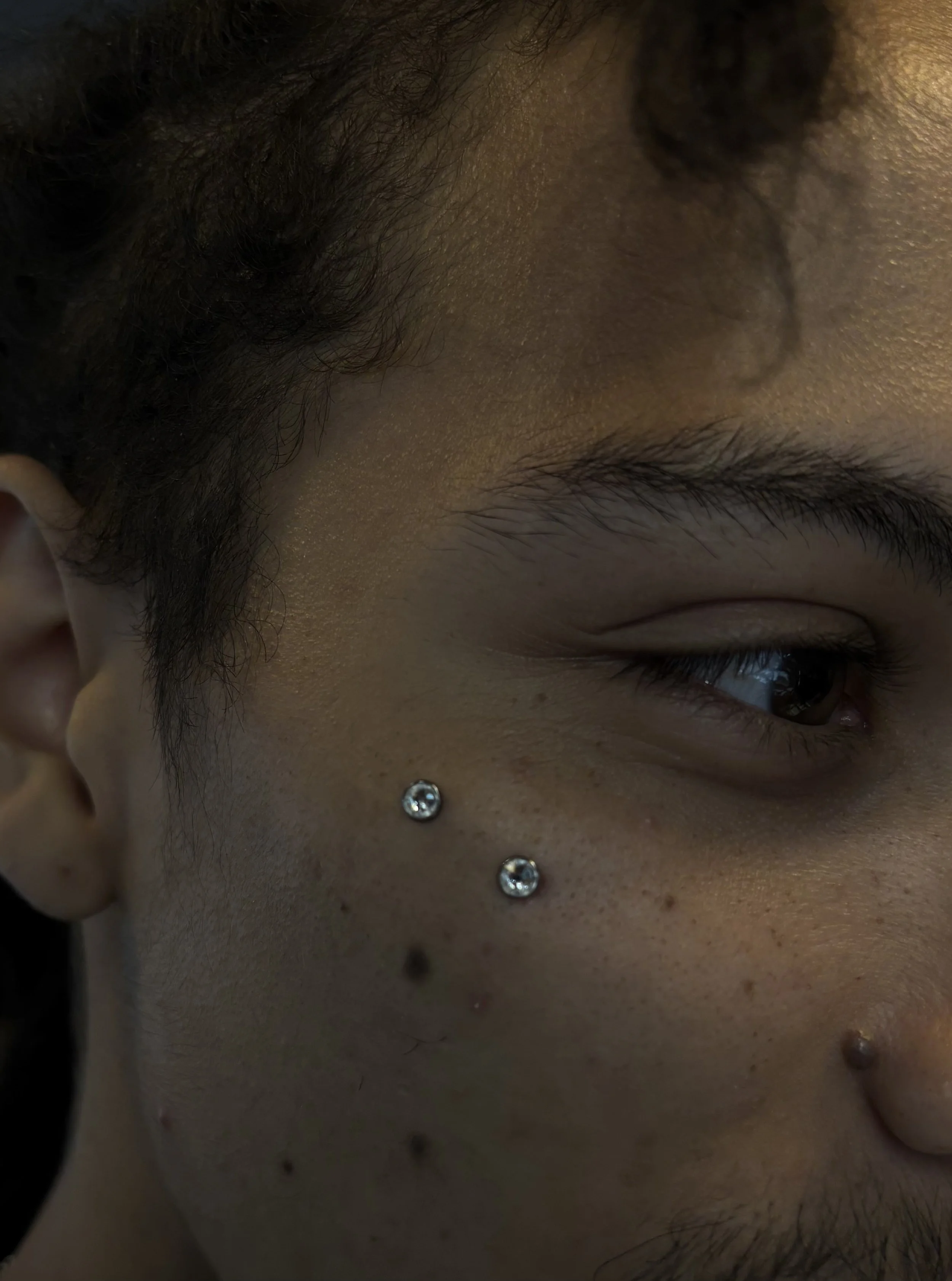 A close-up photo of a person's face focusing on the right eyebrow, upper eye, and cheek, showing jewelry on the face including a nose ring and two piercings below the nose. The person has dark facial hair and smooth skin.