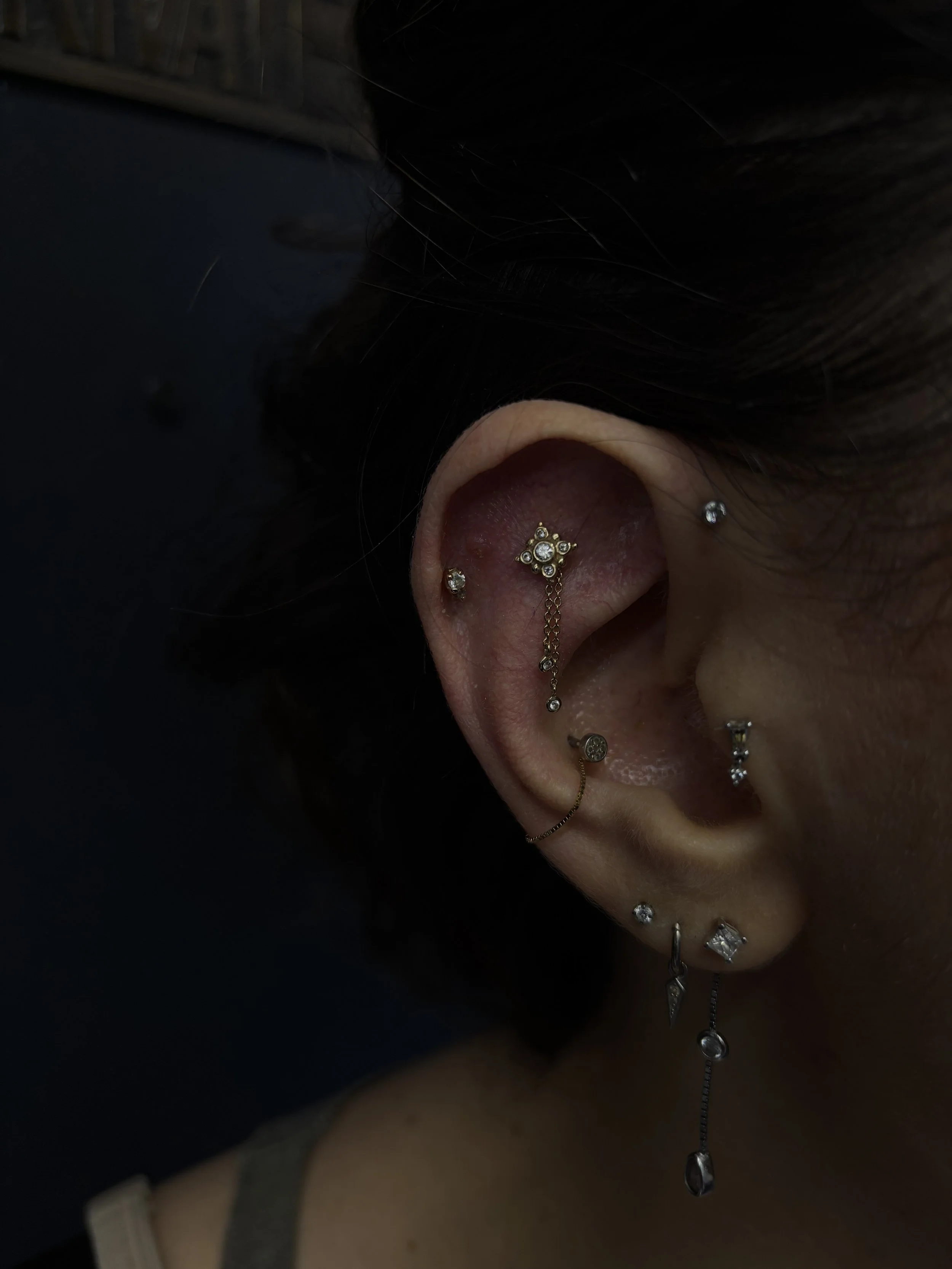 Close-up of a person's ear adorned with multiple earrings, including a decorative chain earring with clear gemstones. The person has dark hair and a dark background.