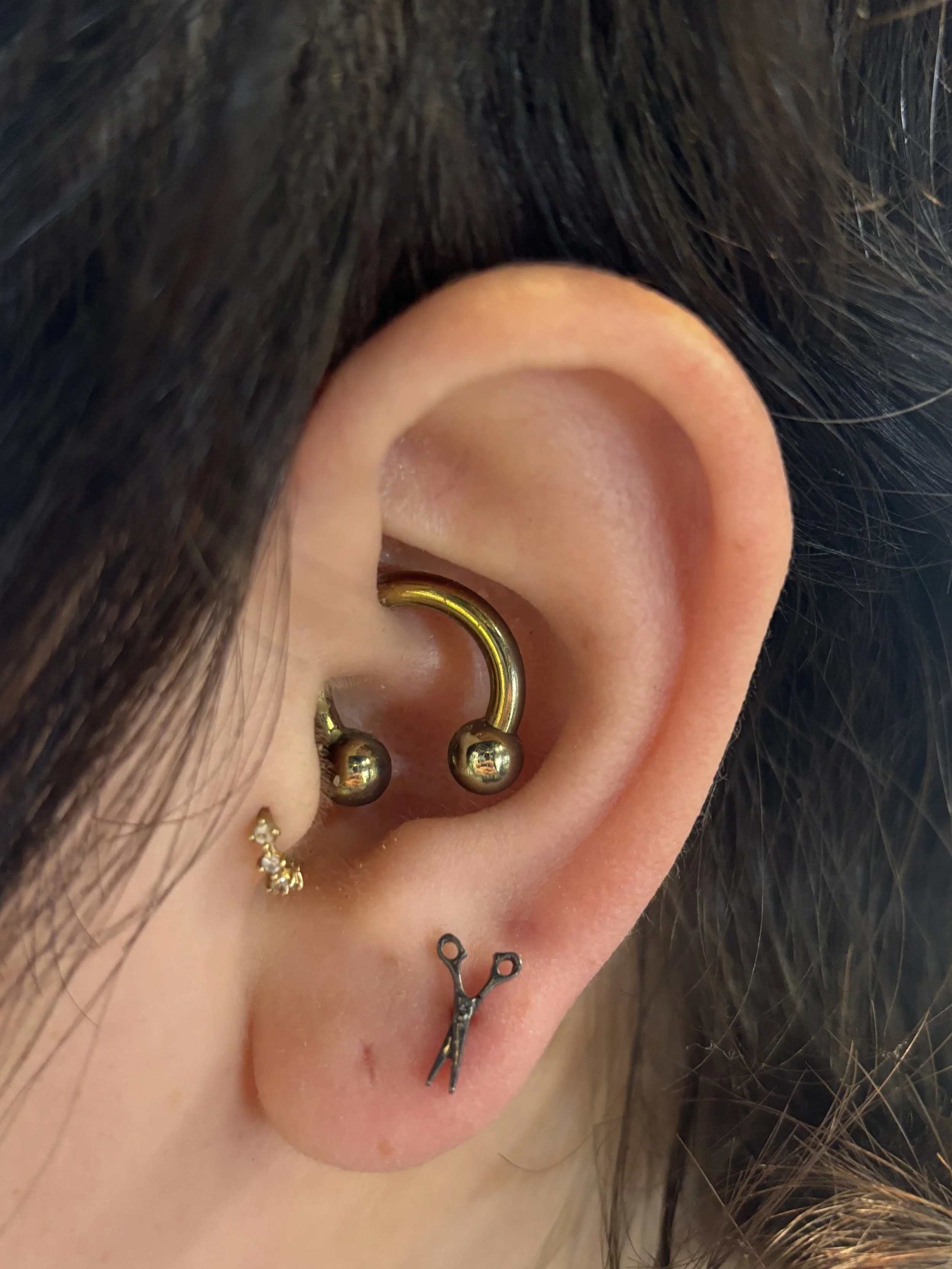 Close-up of a person's ear with multiple piercings, including a gold hoop, a small stud, a shiny ball piercings, and a small decorative design on the lower earlobe. The person has dark hair.