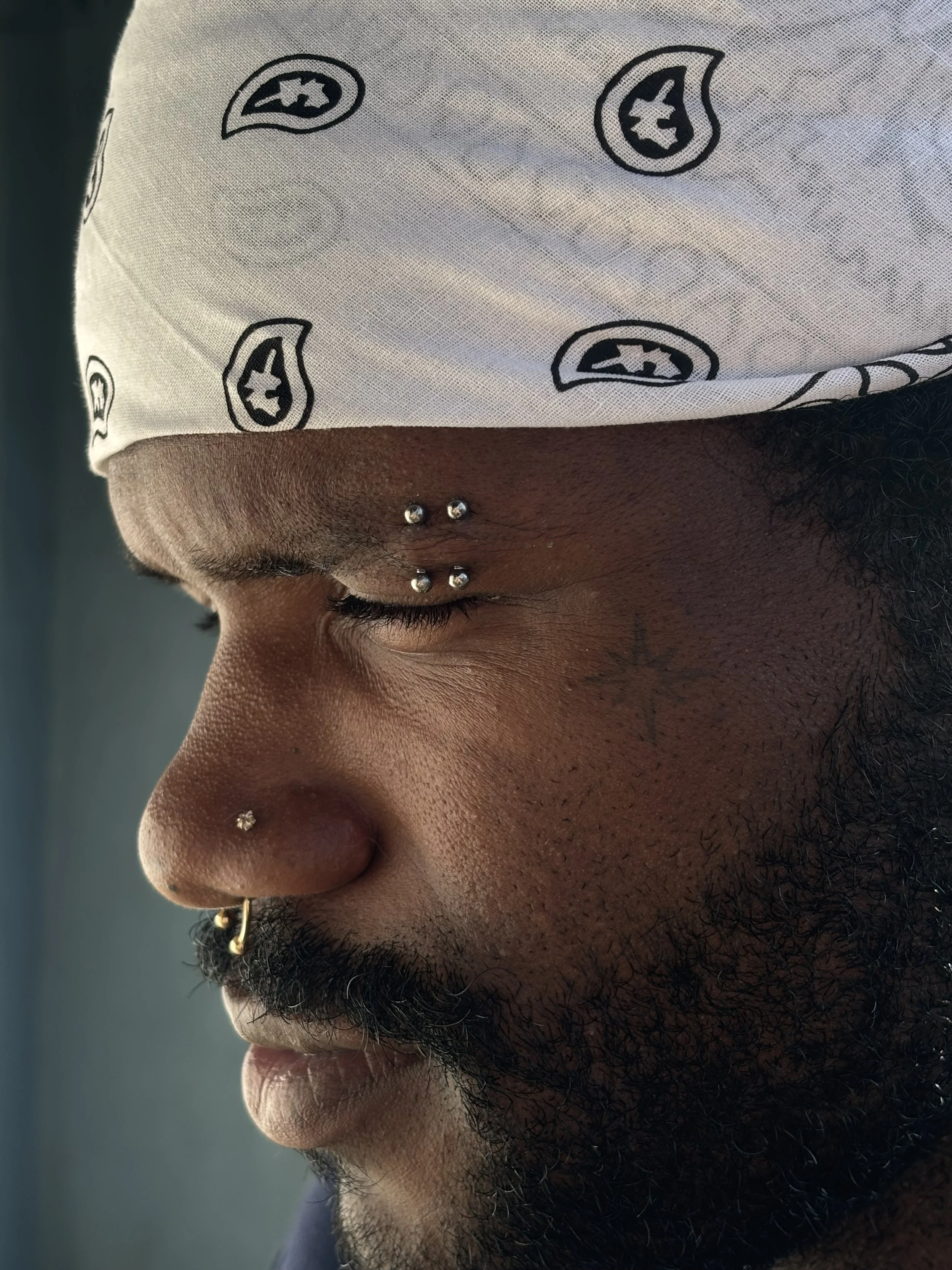 A close-up side profile of a man's face with dark skin, a black beard, and multiple facial piercings including a septum ring, a nostril piercing, and multiple earrings. The man is wearing a white bandana with black paisley prints.