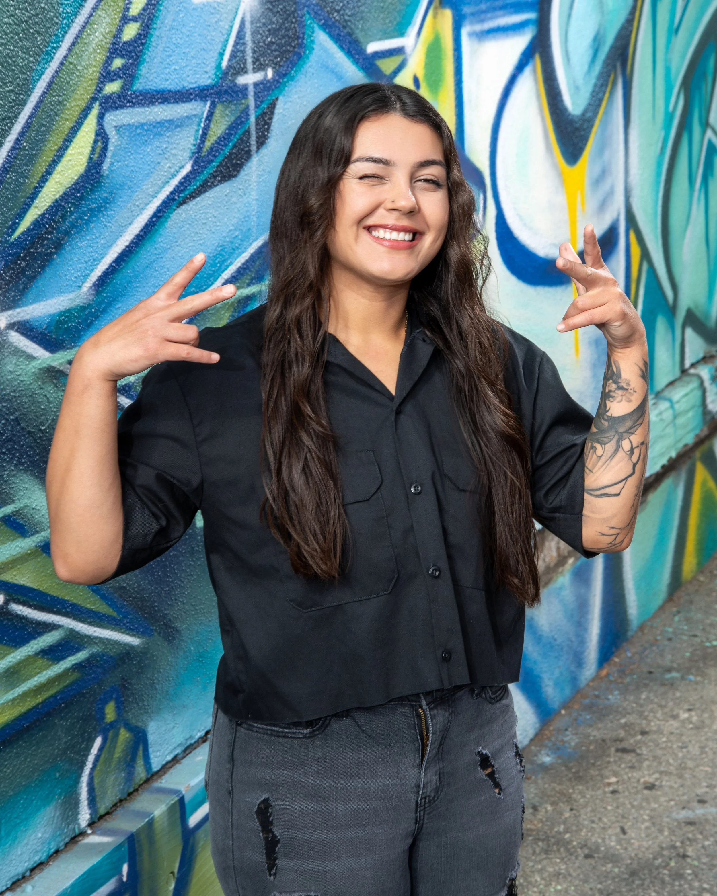 A woman with long dark hair, smiling, making peace signs with both hands, standing in front of a vibrant graffiti wall.