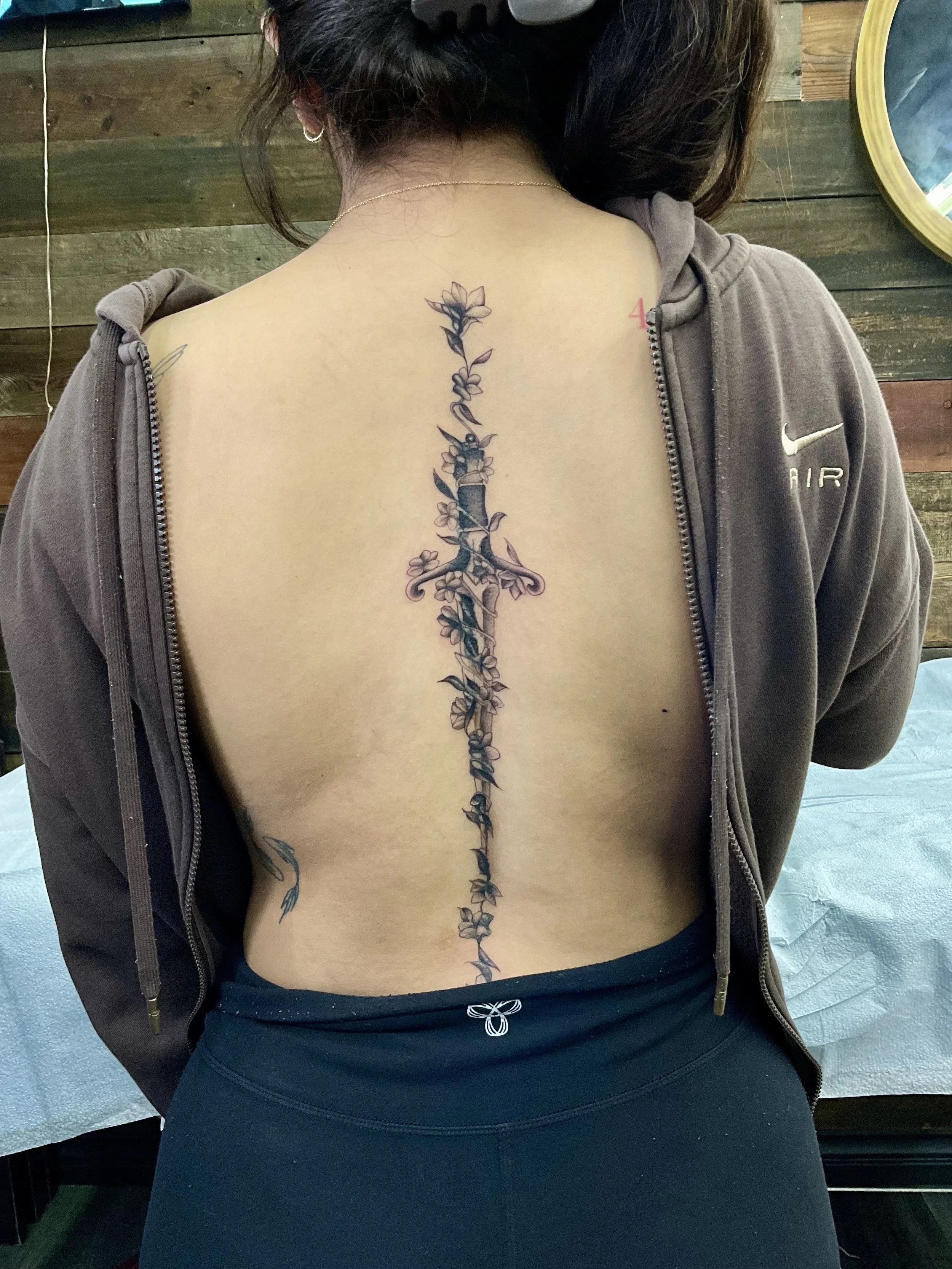 A woman with dark hair wearing a gray Nike zip-up hoodie, black leggings, and a necklace, showing a long floral tattoo running down the middle of her back.