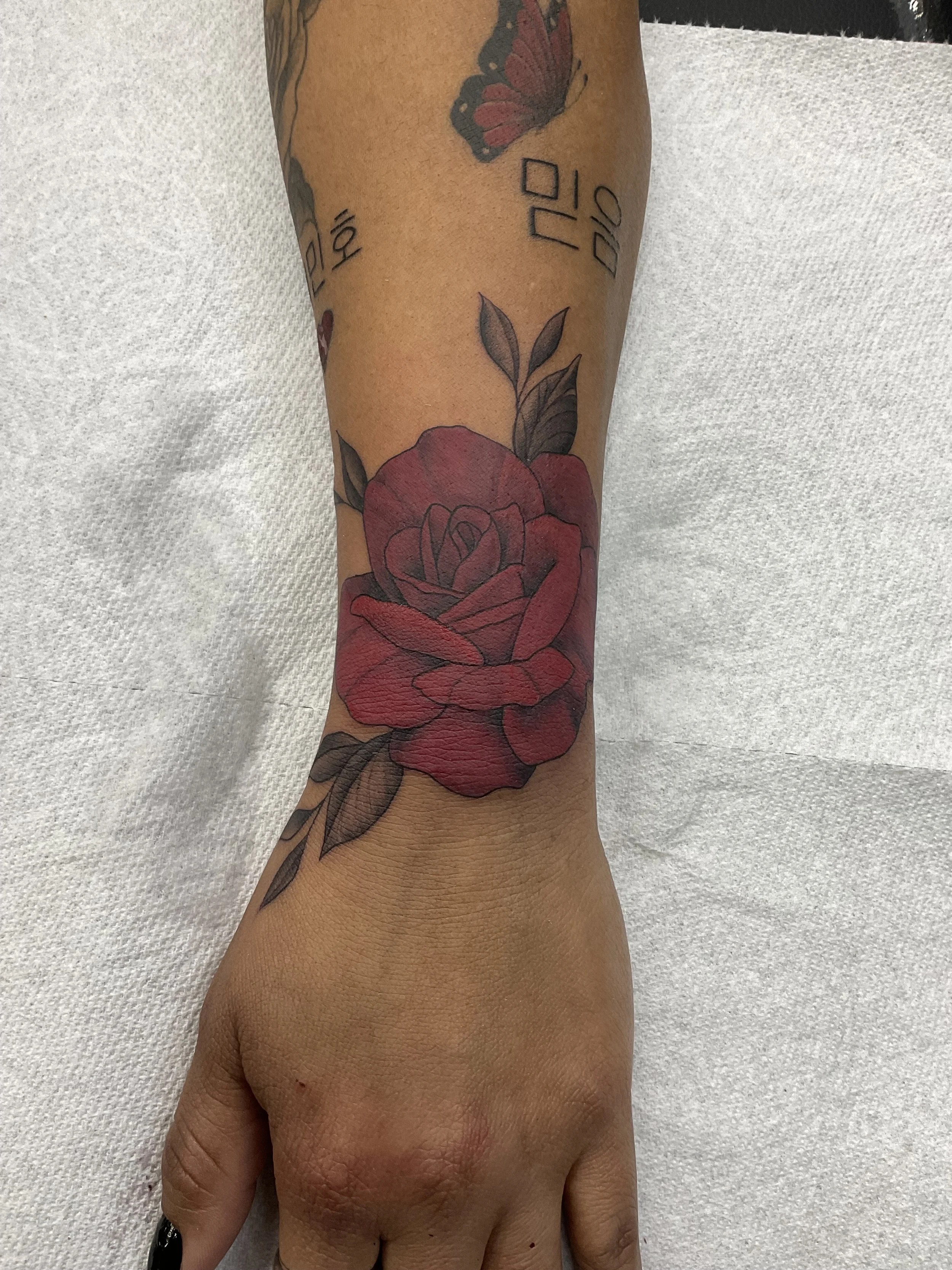 A person's forearm with multiple tattoos, including a large red rose with green leaves, a butterfly, and letters, resting on a white textured surface.