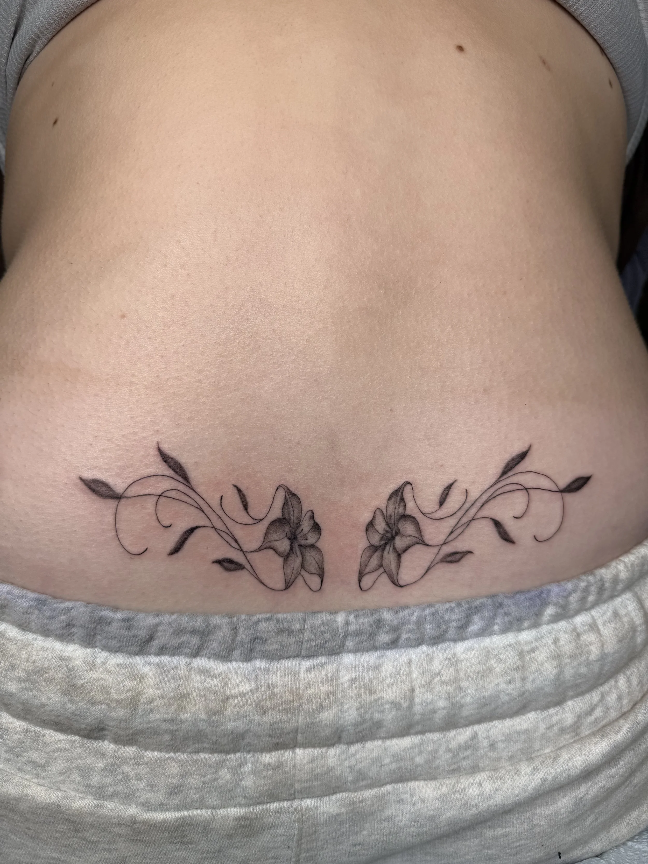 A tattoo of black flowers and vines on the lower stomach or hip area of a person with light skin and a gray shirt.