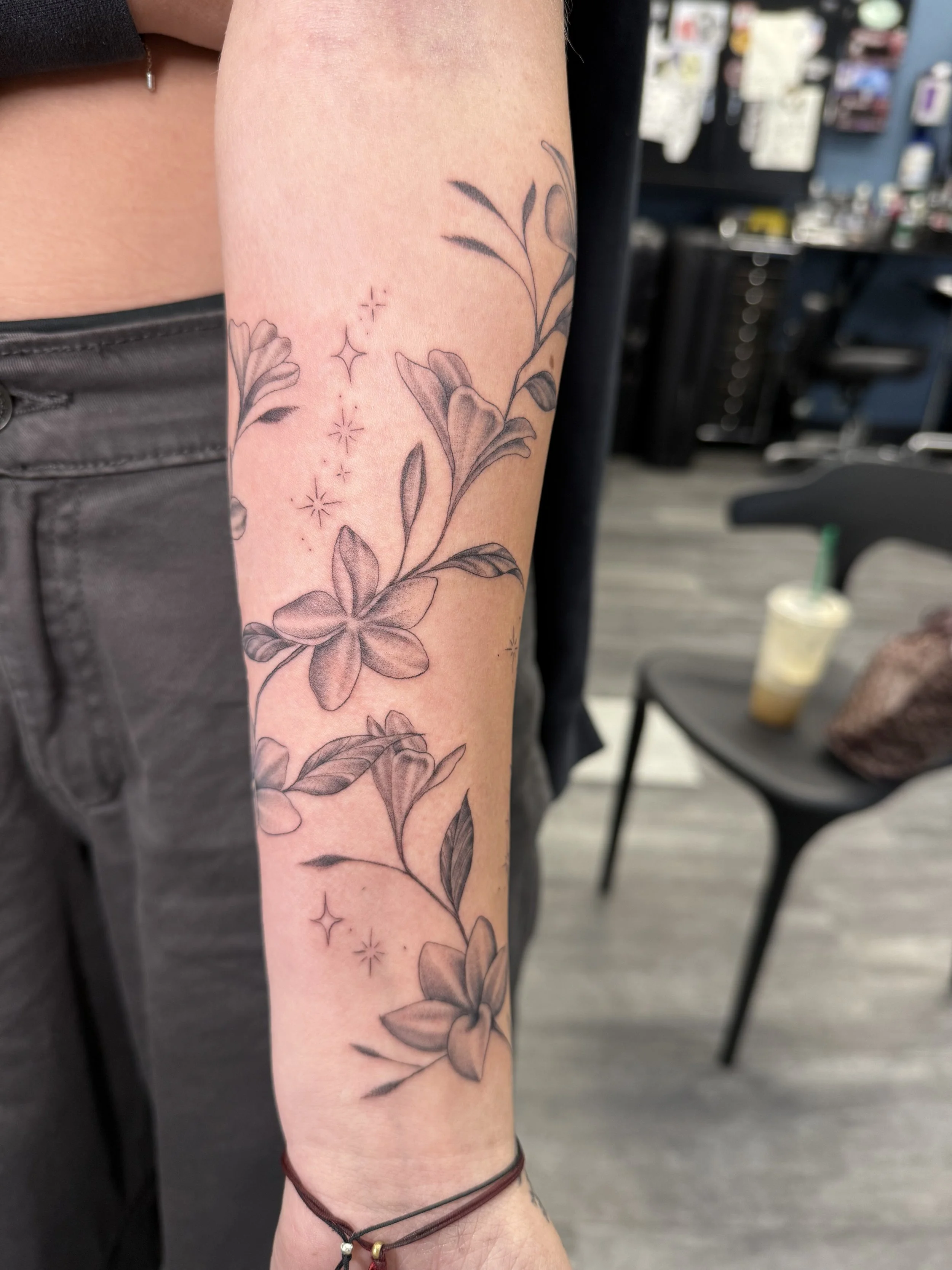 A person's arm with a black ink floral tattoo and small star-like designs, visible in a tattoo parlor setting.