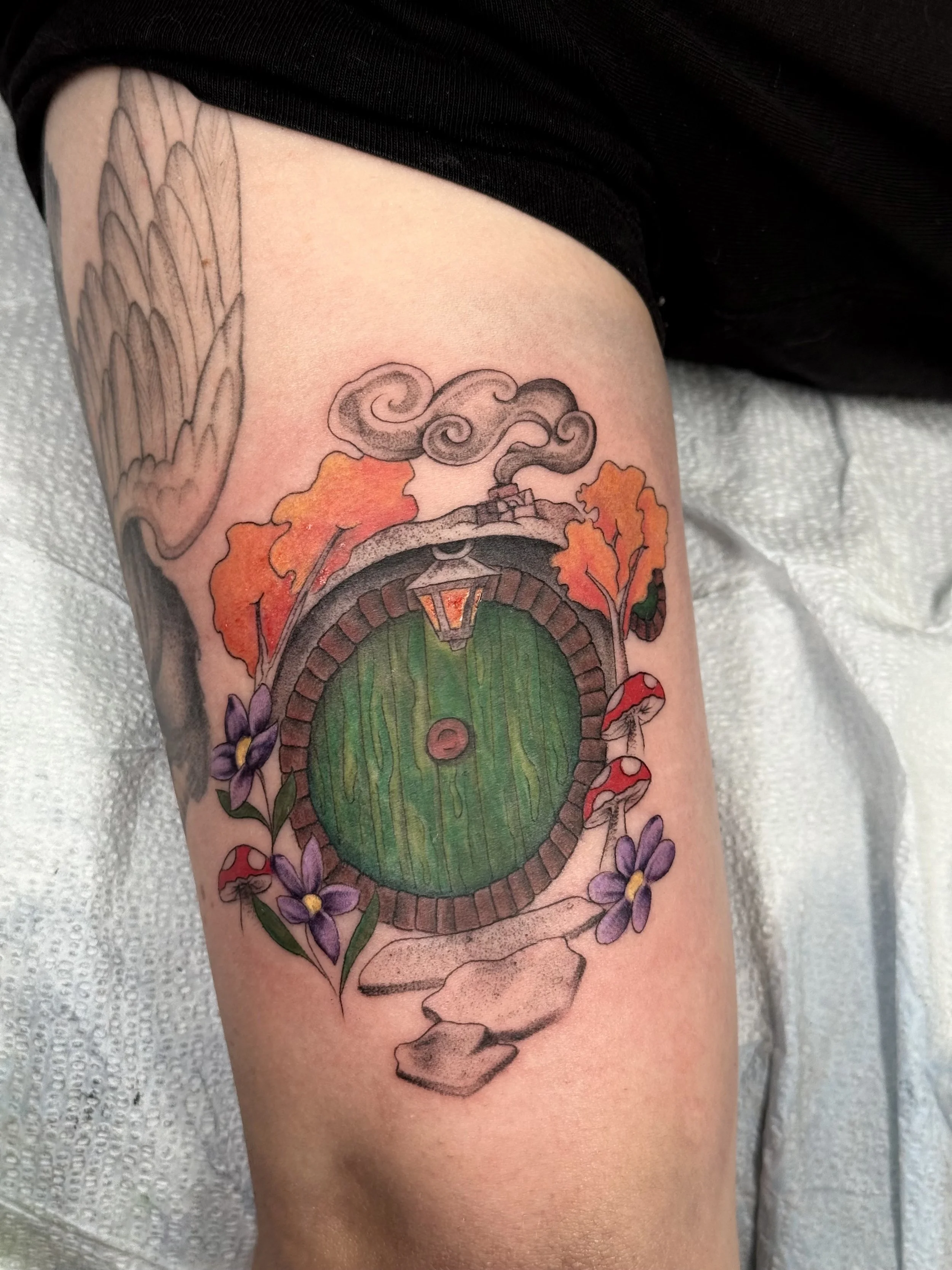 Tattoo of a wooden door with a lantern, surrounded by orange autumn leaves and purple flowers, on a person's upper arm.