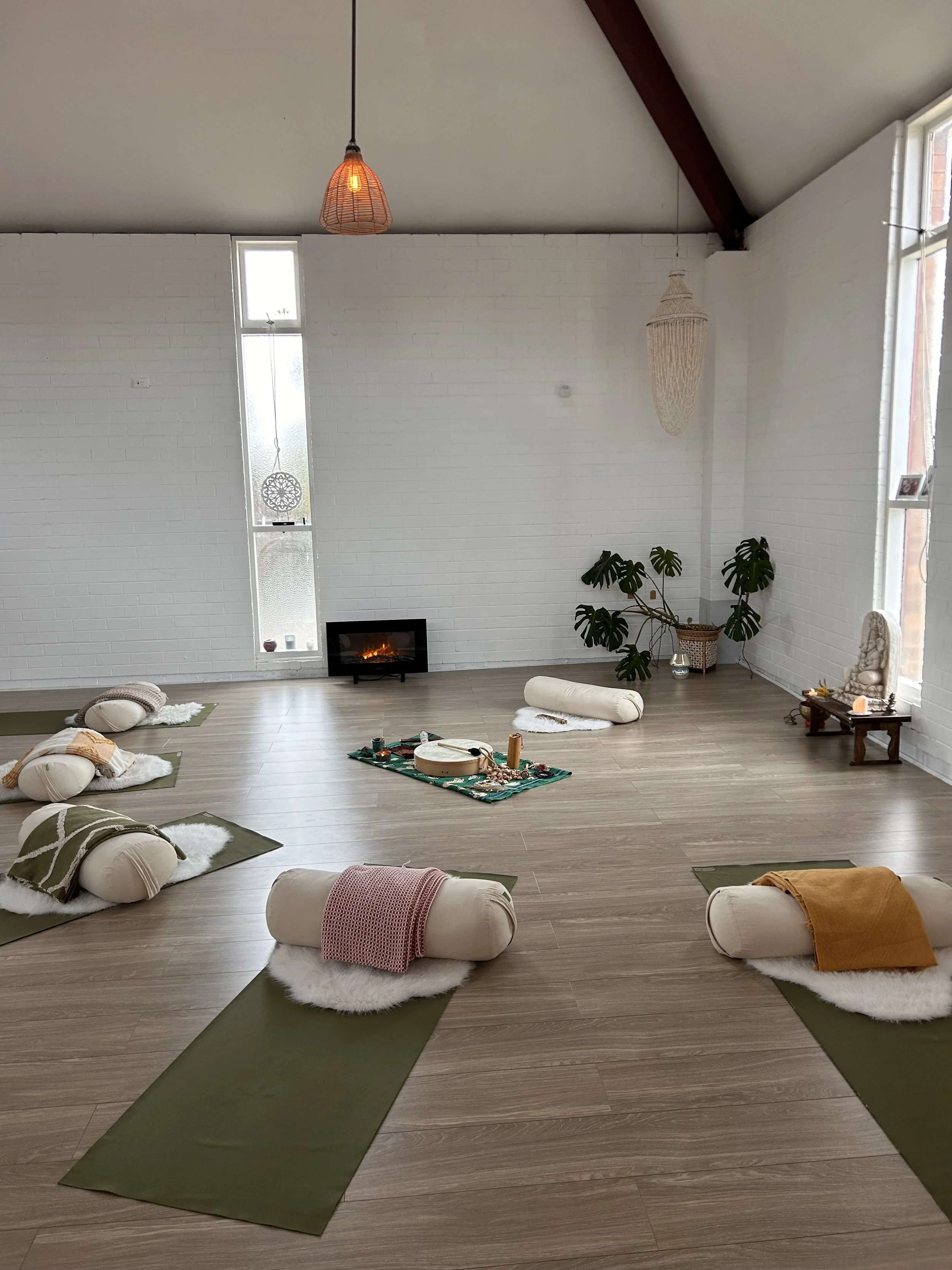 A spacious room with white brick walls, hardwood floors, and high, sloped ceiling. Several yoga mats are arranged on the floor, each topped with a rolled blanket and a cushion. In the background, a small electric fireplace is on the floor, with a narrow vertical window, a hanging decorative ornament, and a large leafy plant.