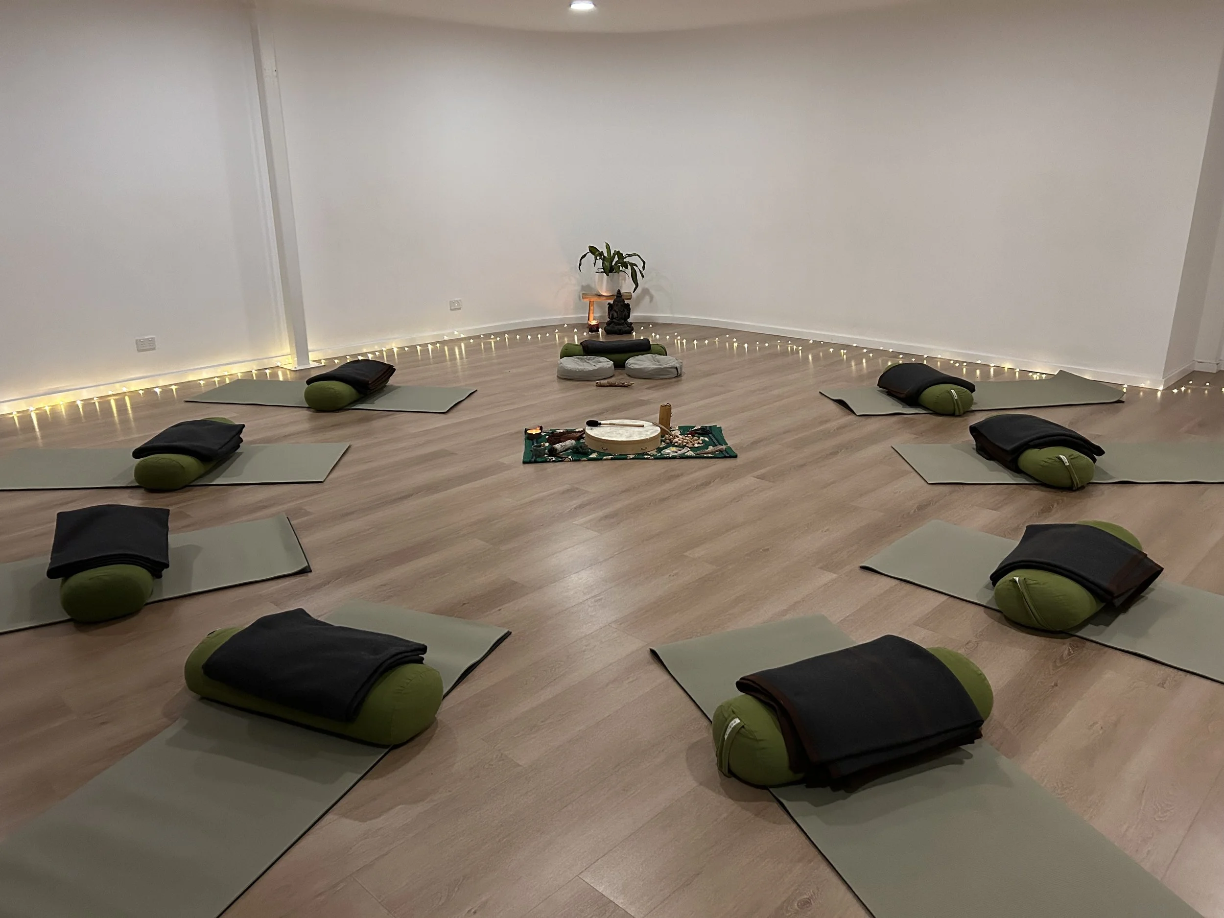 A yoga or meditation class setup with mats, bolsters, and blankets arranged in a semi-circle in a room with wooden flooring and a white wall decorated with a small Buddhist altar and fairy lights