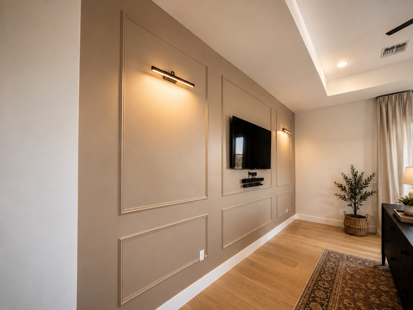 Living room wall with beige paint, decorative trim, mounted flat-screen TV, two wall lights, and a potted plant in a basket. Part of a black dresser with a table lamp and curtains on the window are also visible.