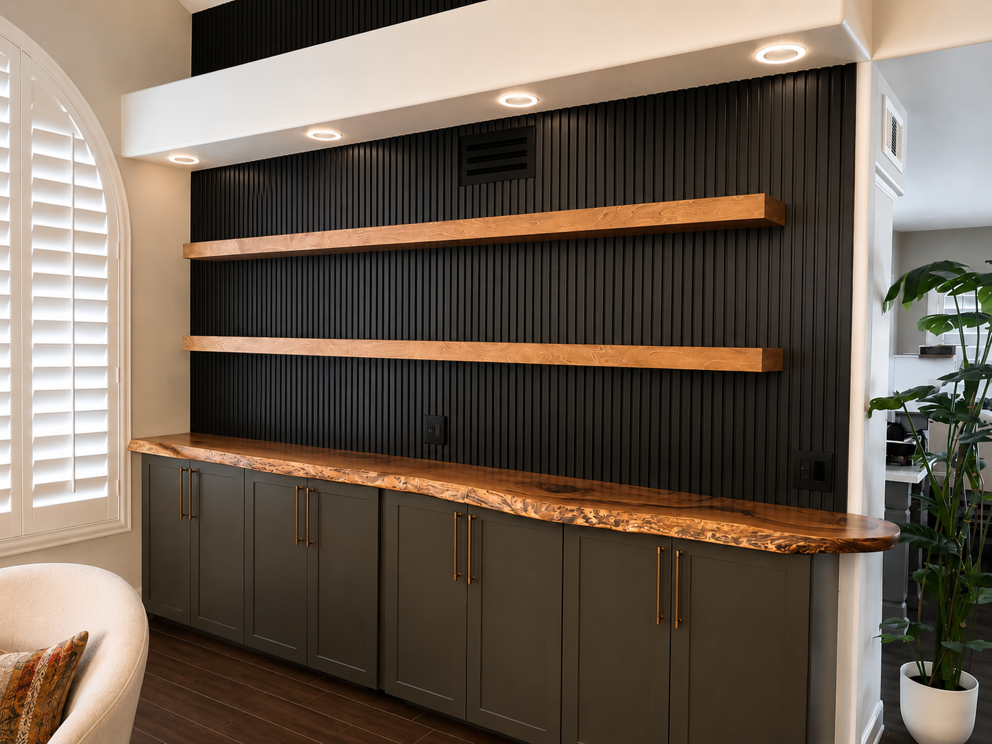 A built-in cabinet with green lower cabinets, a wooden countertop, and two floating wooden shelves on a black ribbed wall. There is a window with white shutters on the left and a potted plant on the right.