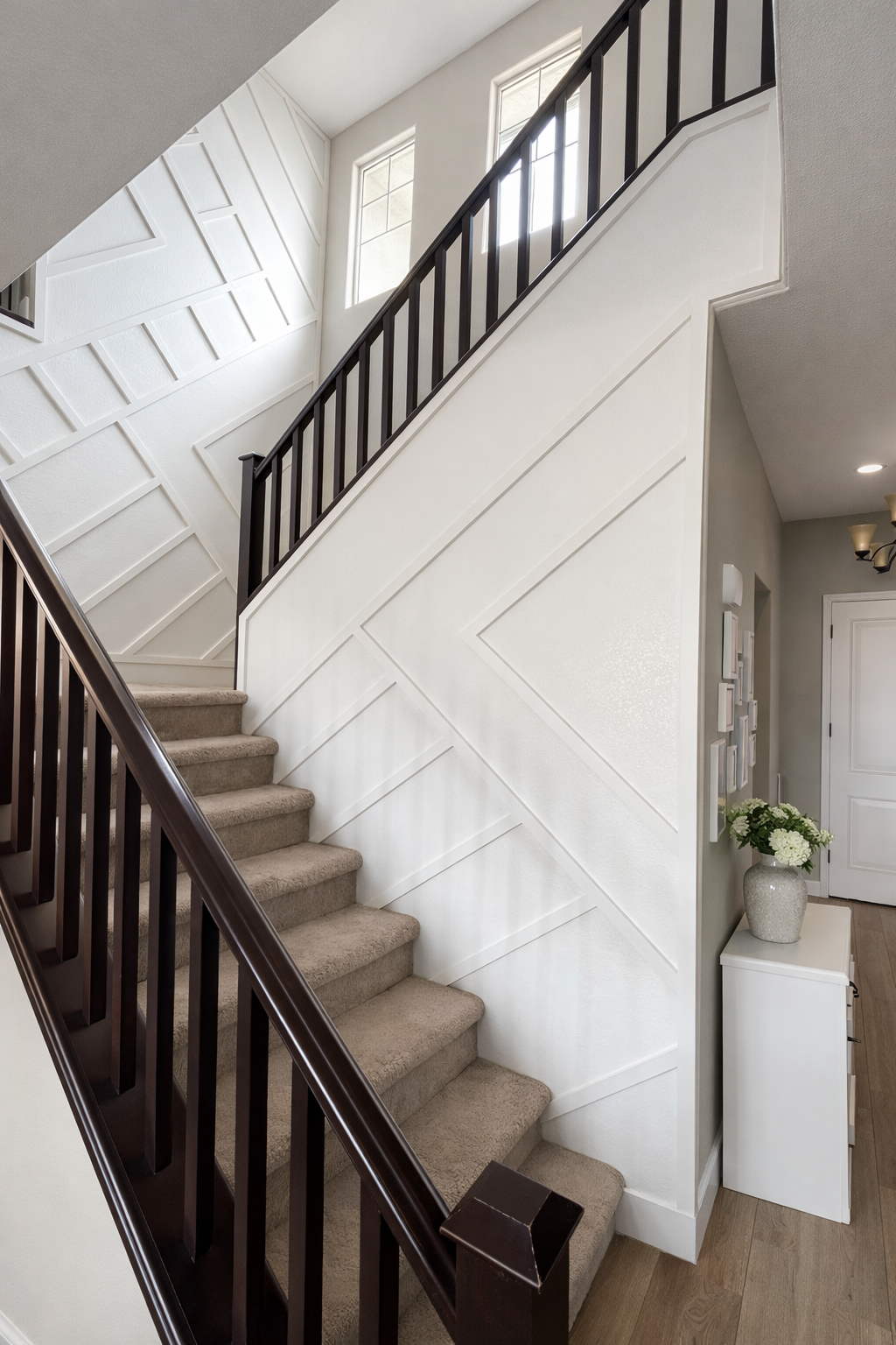Interior of a multi-story house with carpeted staircase, white paneled wall, windows at the top, and a small white table with a flower vase beside a closed door.