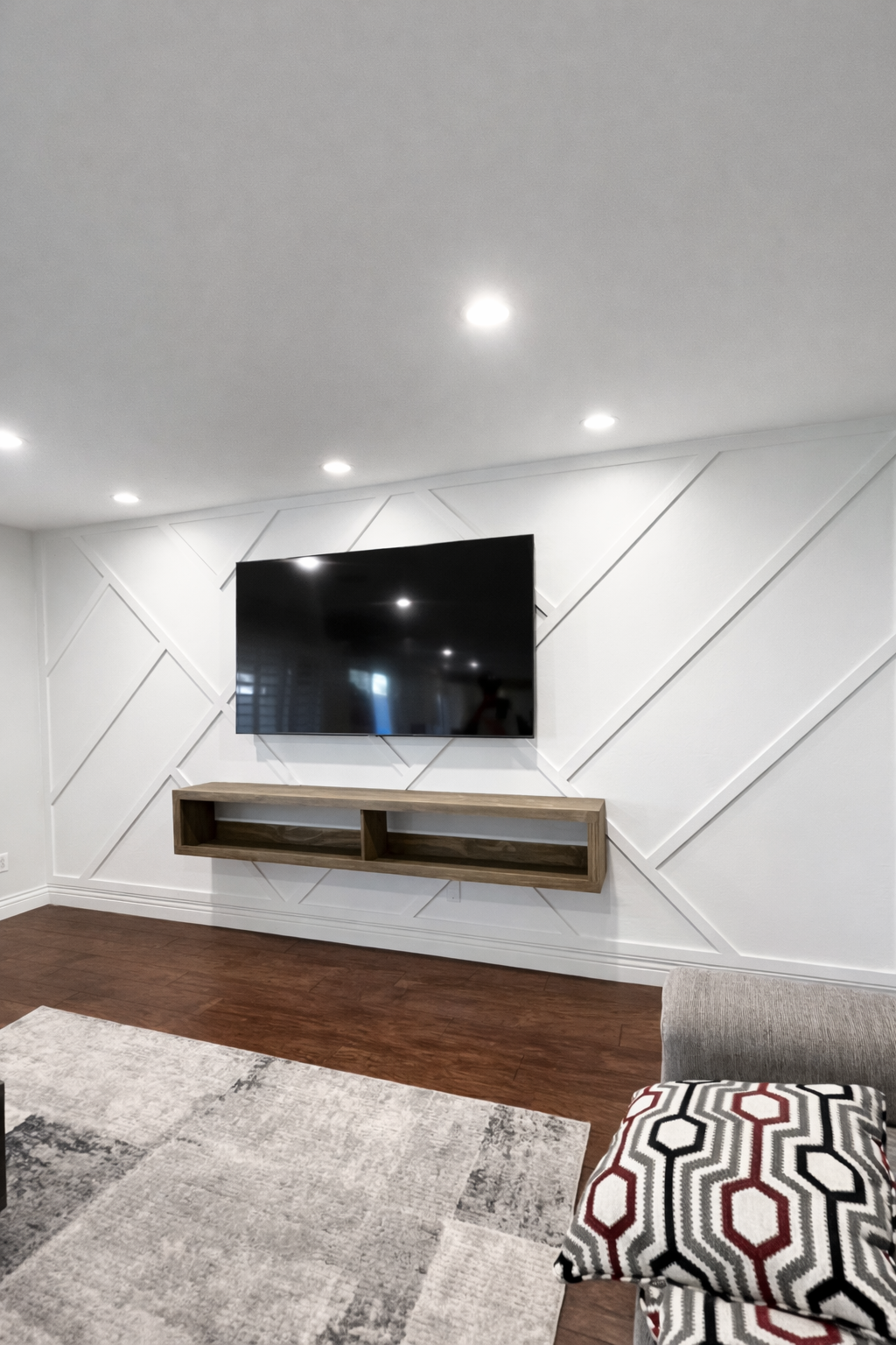 Living room with a wall-mounted flat-screen TV, a wooden floating shelf beneath it, and a patterned cushion on a chair. The room has white walls with geometric wall paneling, a white ceiling with recessed lights, and a wooden floor.
