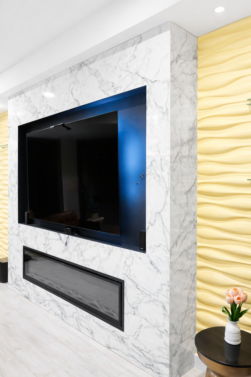 Living room wall with marble finish, mounted flat-screen TV, and a modern fireplace below. To the right, a yellow textured wall with a small black table holding a white vase with pink flowers.
