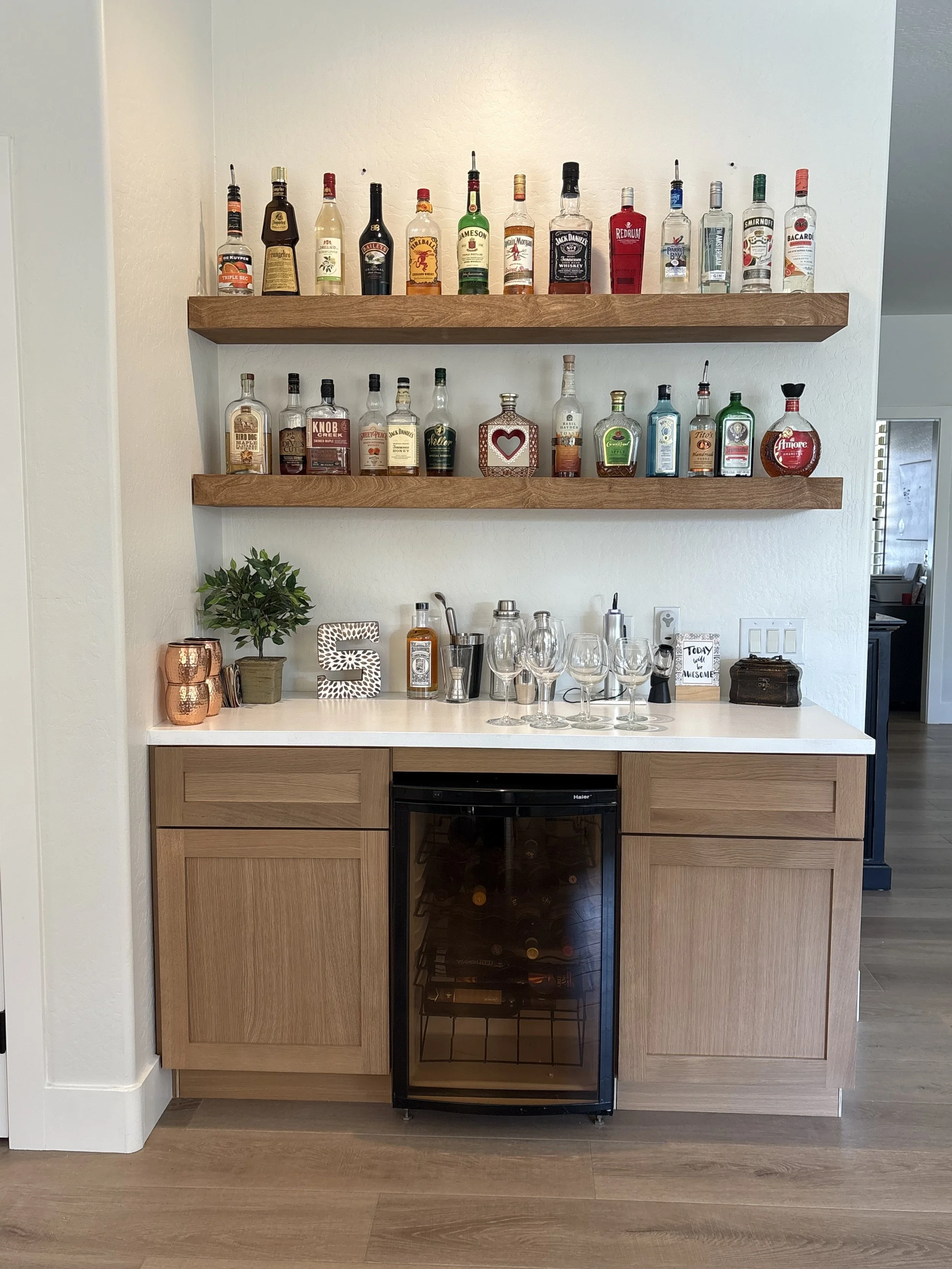 A wooden bar cabinet with a built-in wine cooler, two upper shelves holding various liquor bottles, and a countertop with glasses, a small plant, and decorative items.