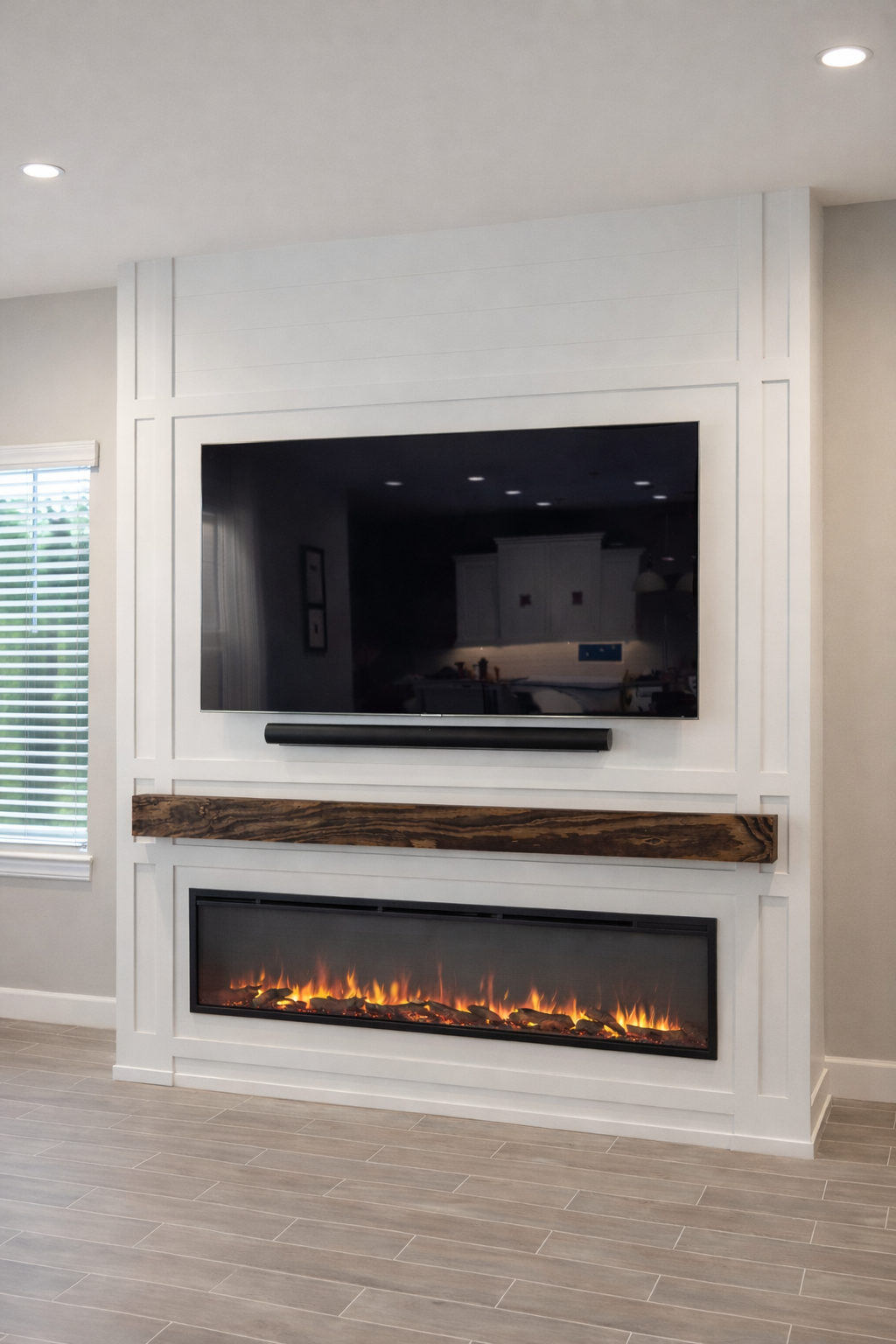A modern living room with a flat-screen TV mounted on a white wall, above a linear electric fireplace with flames, a wooden mantel, and a large window with blinds.