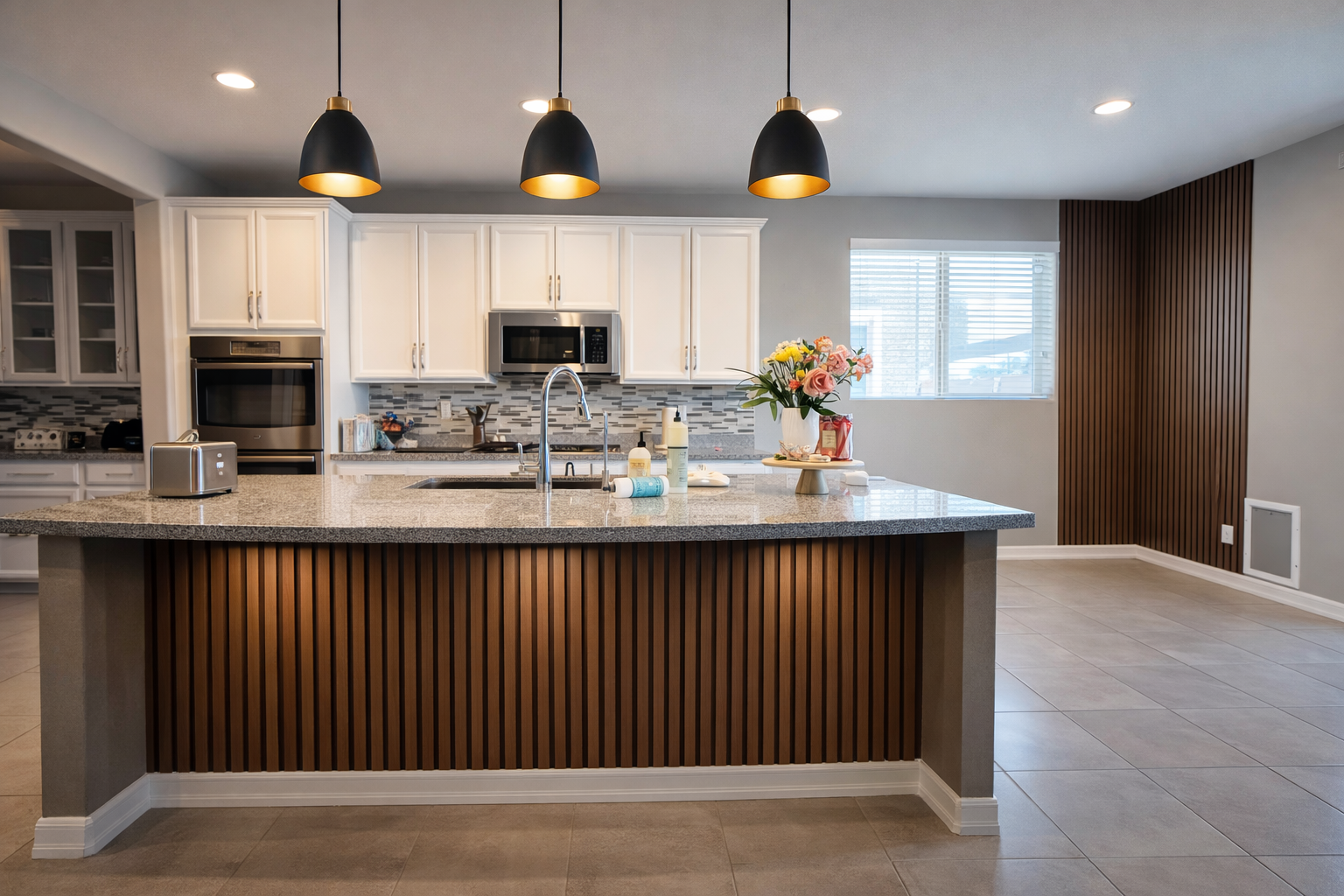 Modern kitchen with white cabinets, a granite island with wood paneling, black pendant lights, stainless steel appliances, a tiled backsplash, a window with blinds, and a vase of flowers on the island.