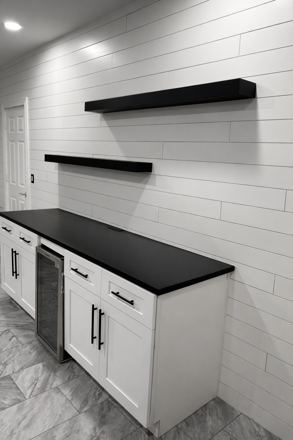 Modern white kitchen cabinet with black countertop, black handles, and two black floating shelves mounted on a wood-paneled wall with gray marble floor.