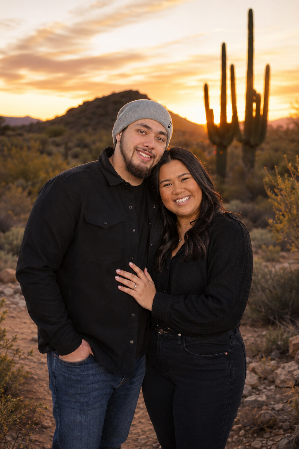 A smiling couple stands close together in a desert landscape with a large saguaro cactus and mountains in the background at sunset.