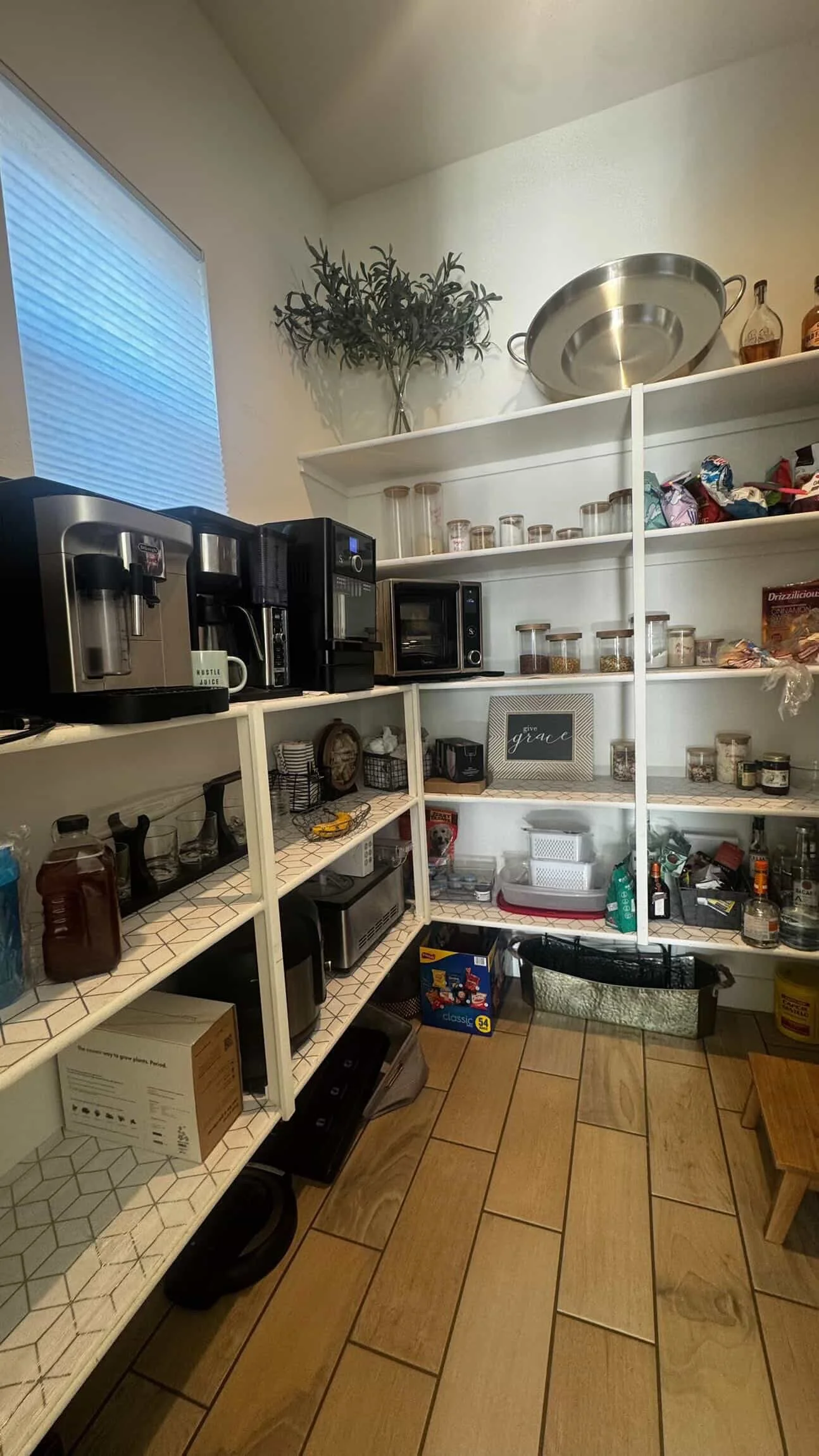 A kitchen pantry with white shelving stocked with jars of spices, snacks, and kitchen appliances including a coffee maker, microwave, and toaster oven. There are cabinets and a small table or island below, with various kitchen items, boxes, and utensils.