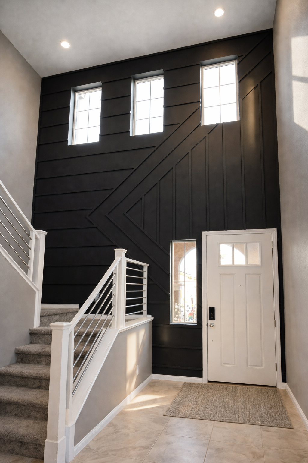 Modern entryway with a black accent wall featuring geometric paneling, three vertical windows, a white front door with a window, a small window next to it, tiled floor, and staircase with white and metal railing.
