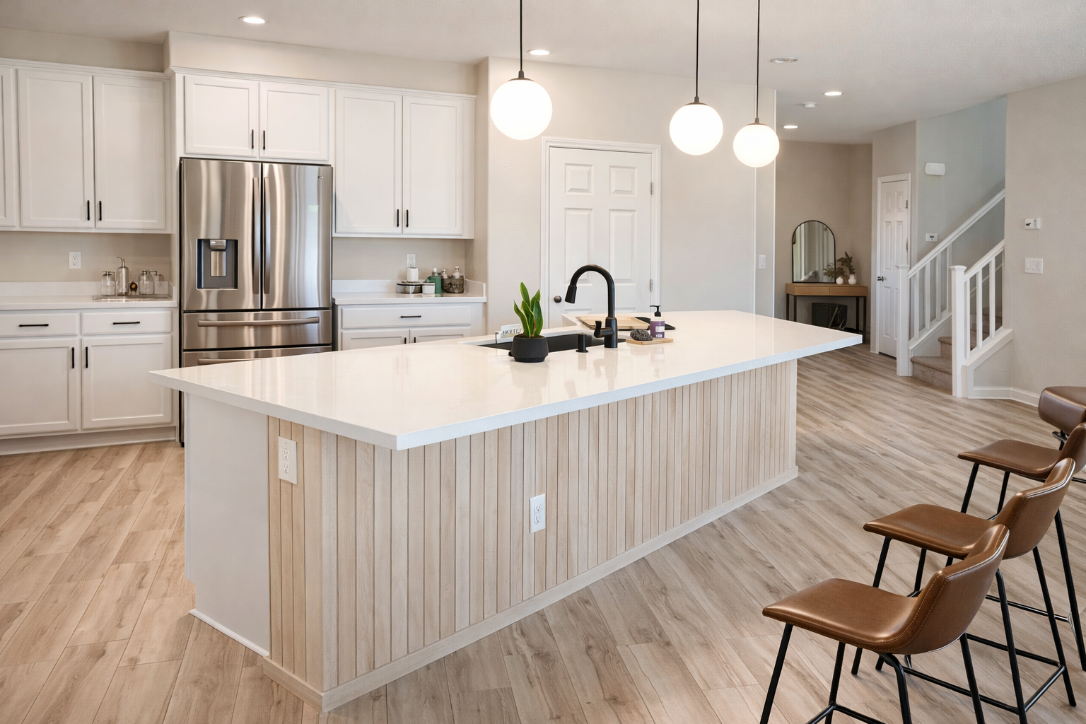Modern kitchen with white cabinets, stainless steel refrigerator, a large island with a white countertop, three brown chairs, pendant lights, and a wooden floor.