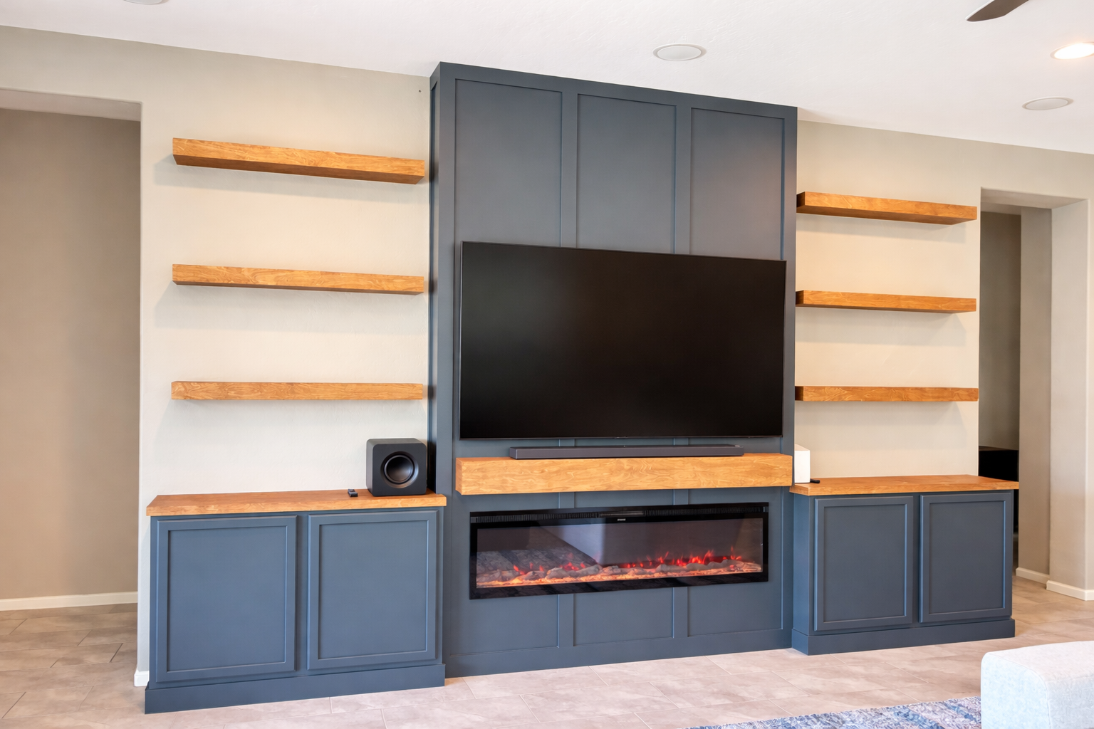 Living room entertainment wall with a large flat-screen TV mounted above an electric fireplace, surrounded by blue cabinetry and wooden shelves.