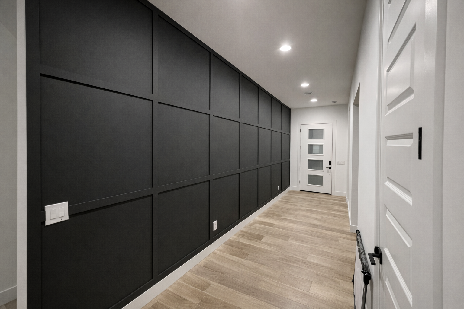 Hallway with a black paneled accent wall, white walls, a white front door with four rectangular windows, and wooden flooring.