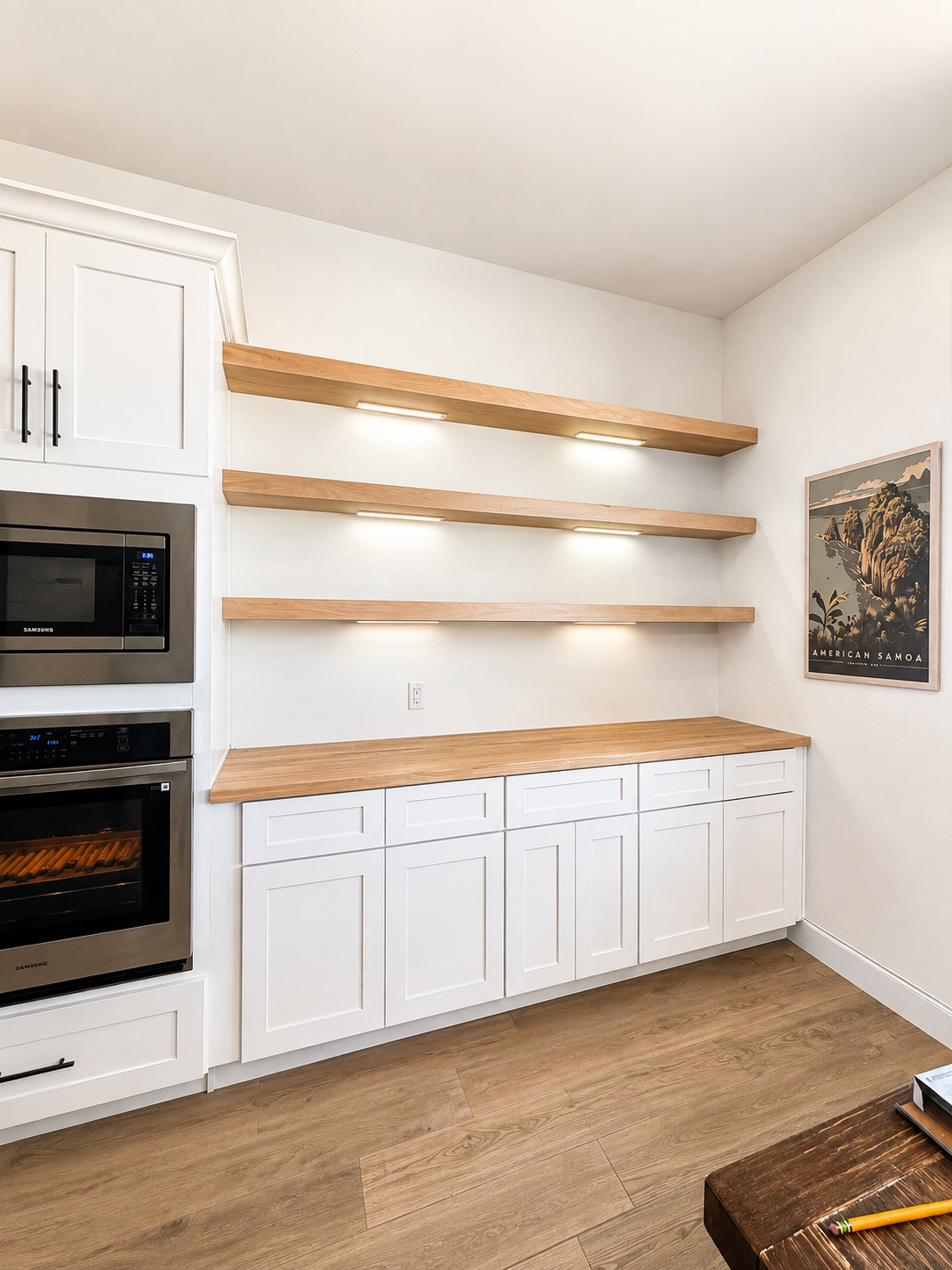 Empty kitchen with white cabinets, wooden countertop, and open wooden shelves with built-in lighting, tropical themed framed poster on the wall.