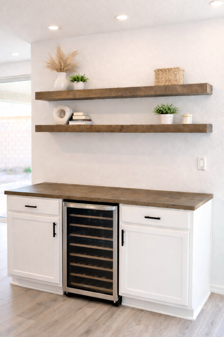 Kitchen corner with white cabinets, a wine cooler, wooden countertop, and two floating wooden shelves decorated with plants, books, and a basket.