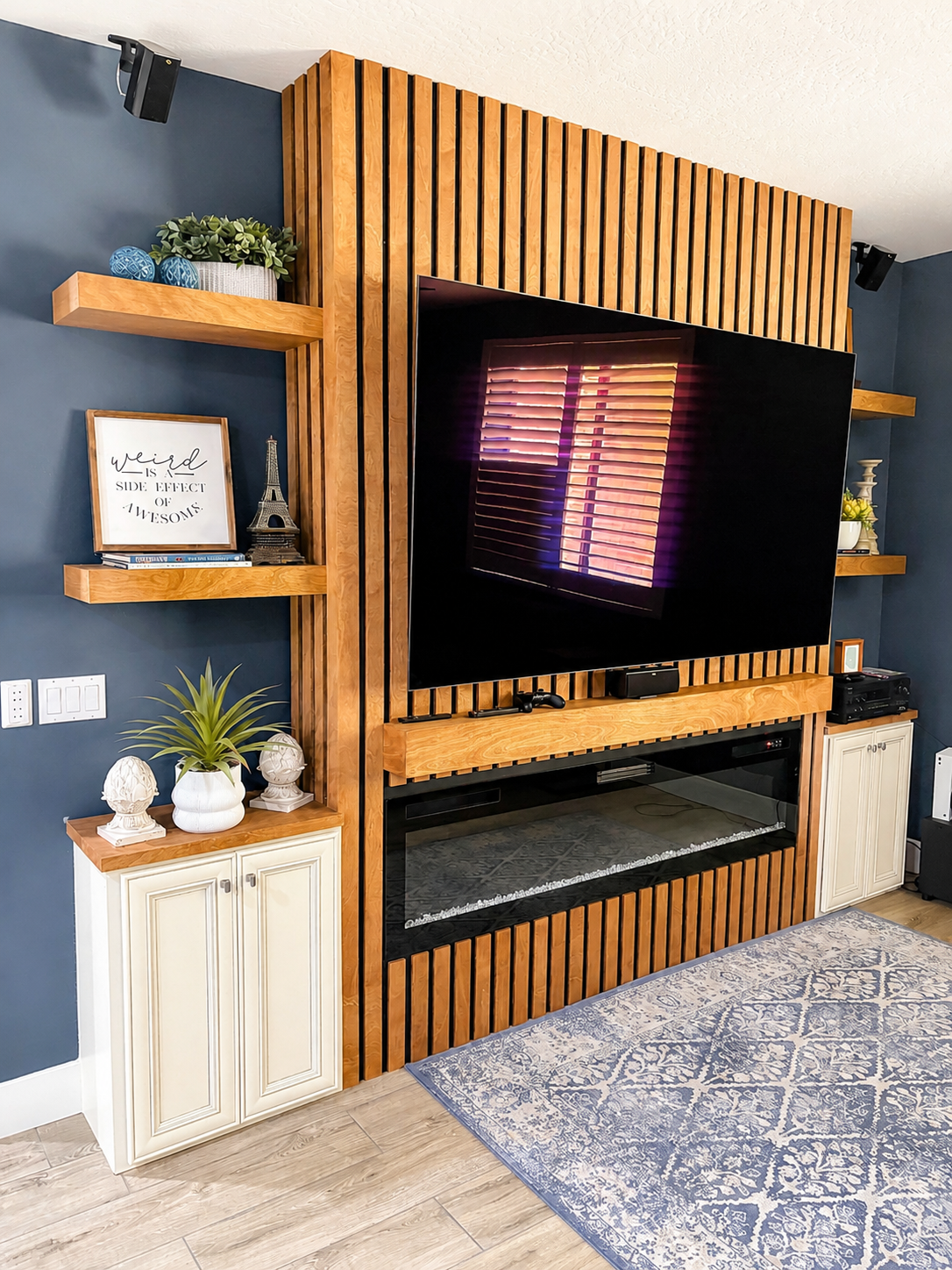 Living room entertainment area with a large flat-screen TV mounted on a wooden slat wall, surrounded by blue walls, wooden shelves with decorative items, a white cabinet with potted plants, and a patterned rug on a wooden floor.