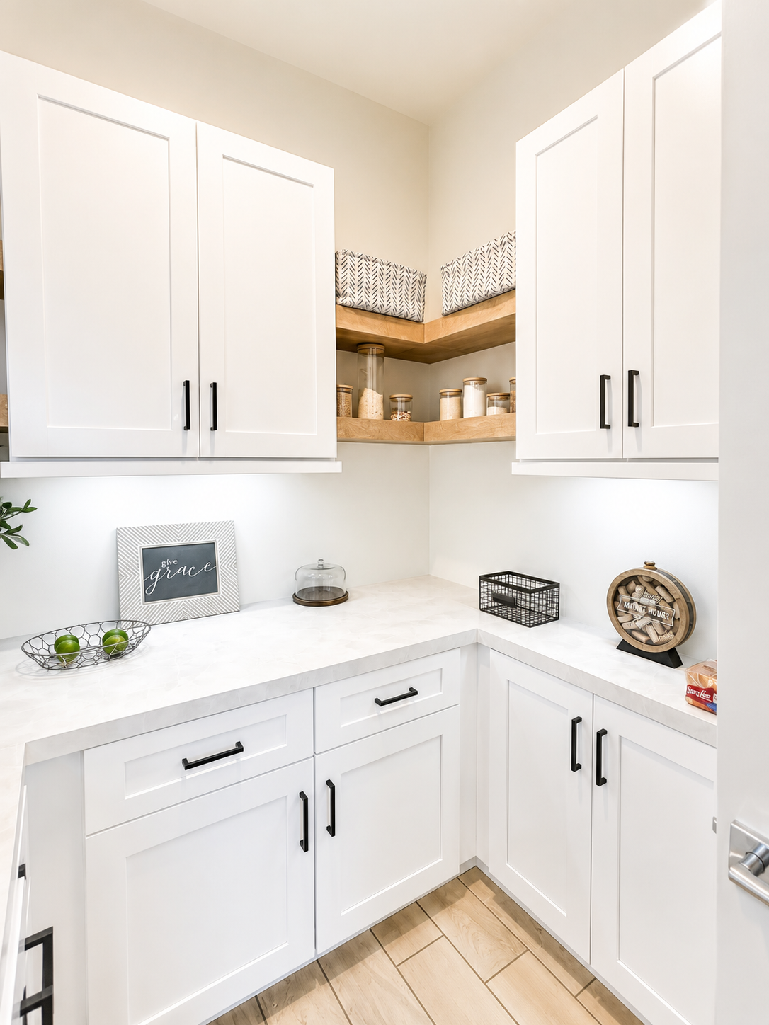 White kitchen corner with black hardware, open wooden shelves with jars, decorative sign with 'give grace', small tray with green apples, glass cloche, wire basket, wooden round bread box, and countertop items.