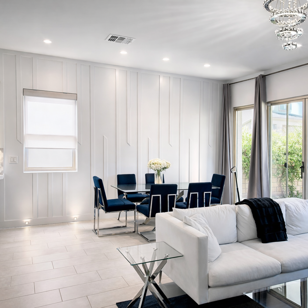 Modern living room with white walls, white tile flooring, and large windows with gray curtains. There is a white sofa with a black throw blanket, a glass side table, a glass dining table with six navy blue chairs, and a chandelier hanging from the ce