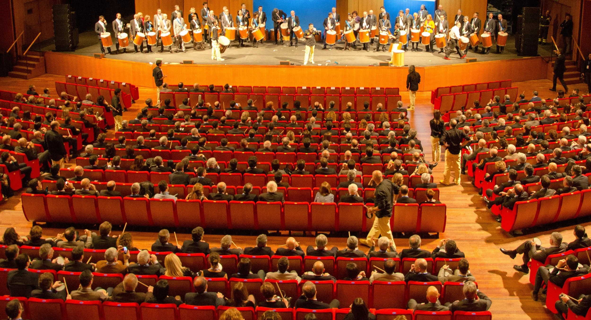 Grande auditorium con un gruppo di persone che suona tamburi sul palco davanti al pubblico.